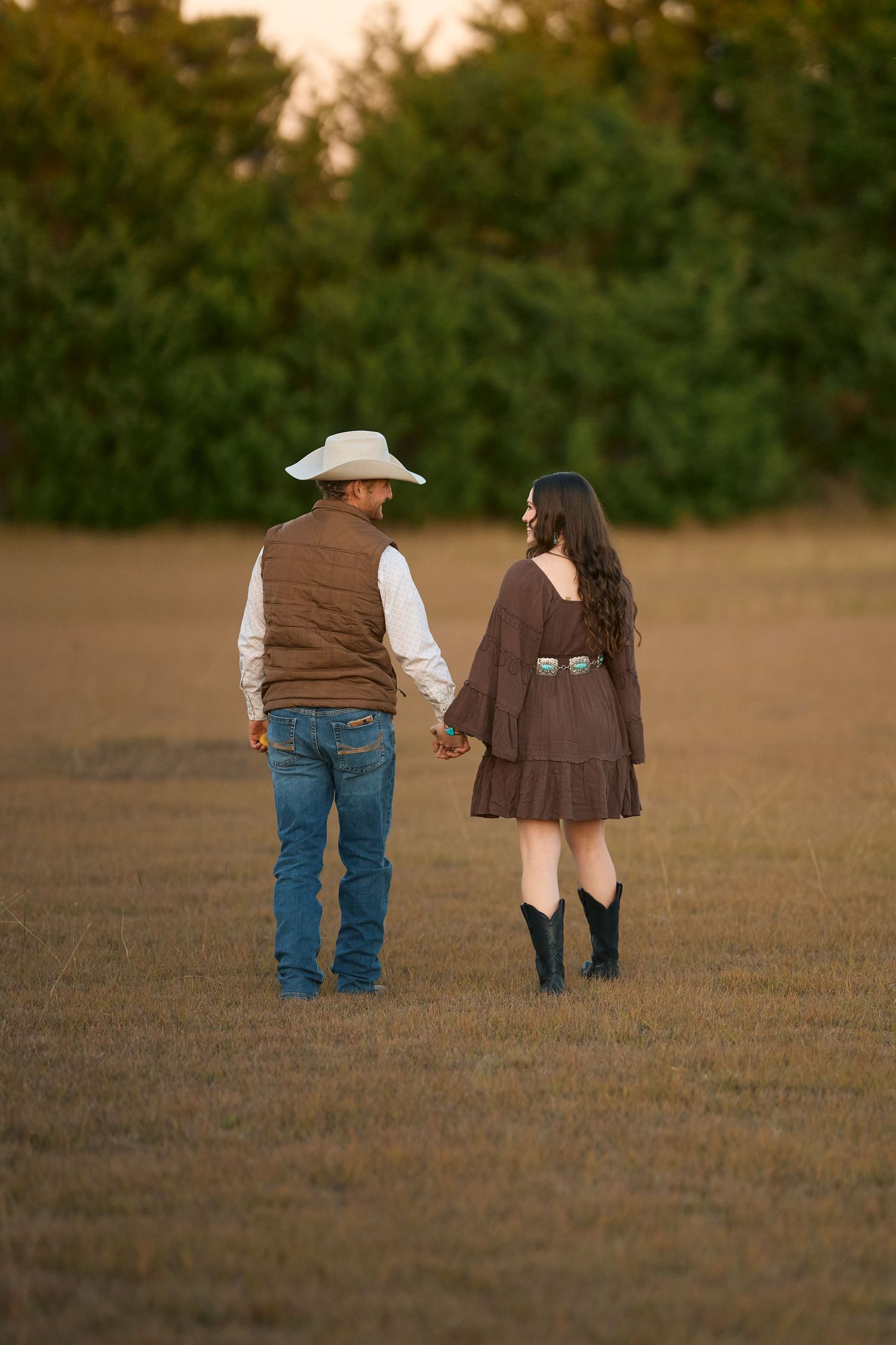 A couple walks together through a brown field at sunset wearing casual western attire.