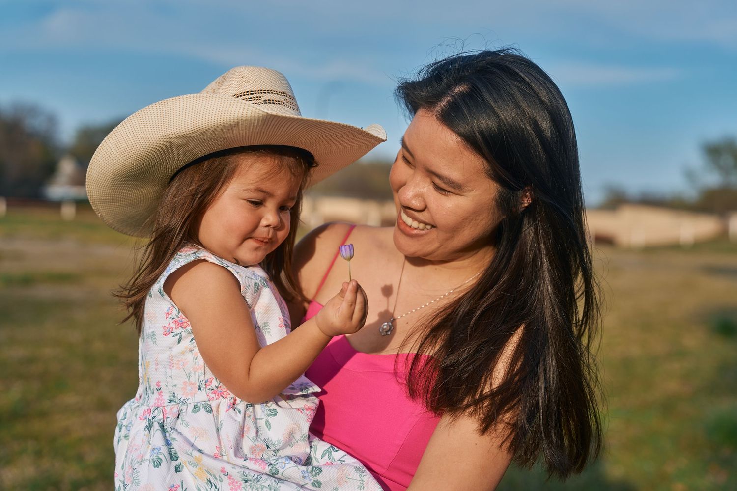 A smiling mother in a pink dress holds her toddler daughter wearing a cowboy hat outdoors on a sunny day.