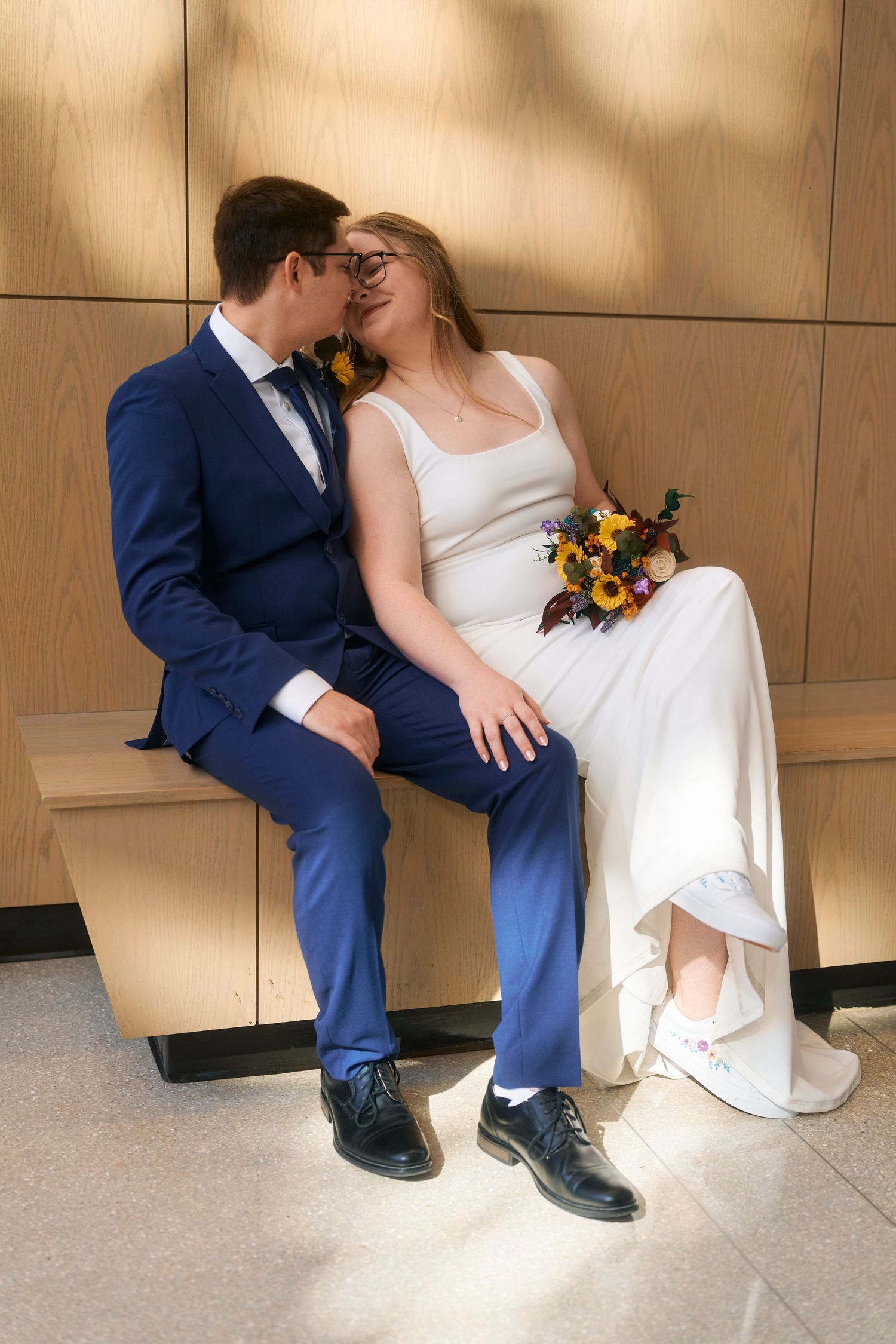 A couple in wedding attire share an intimate moment on a wooden bench against a neutral wall.