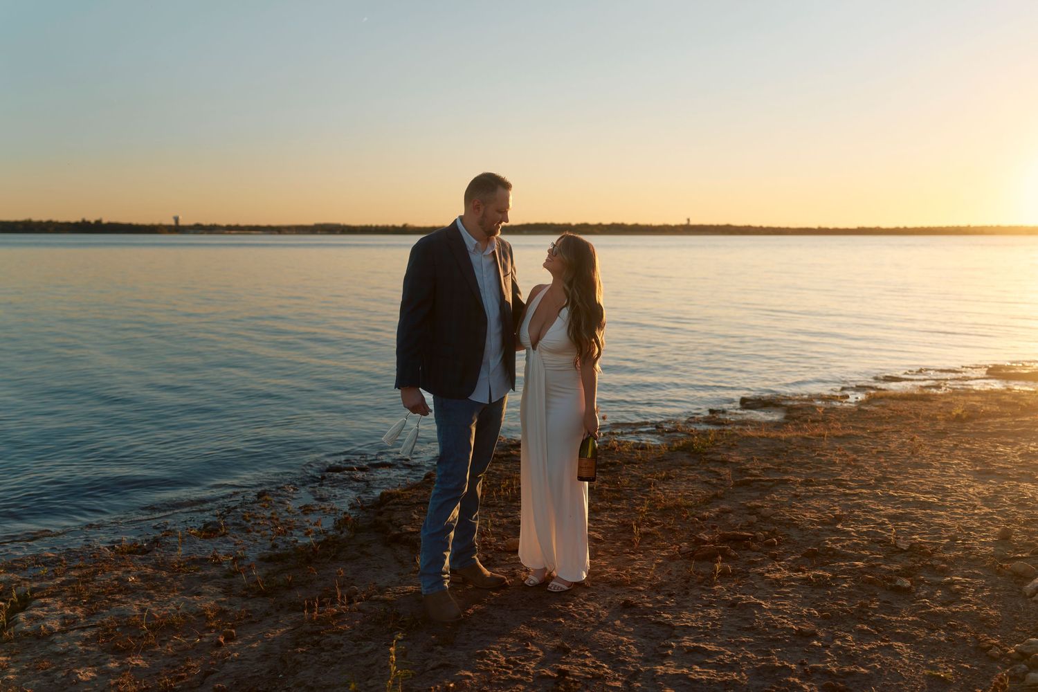 Romantic couple stands together on a beach shoreline at sunset with calm water in the background.