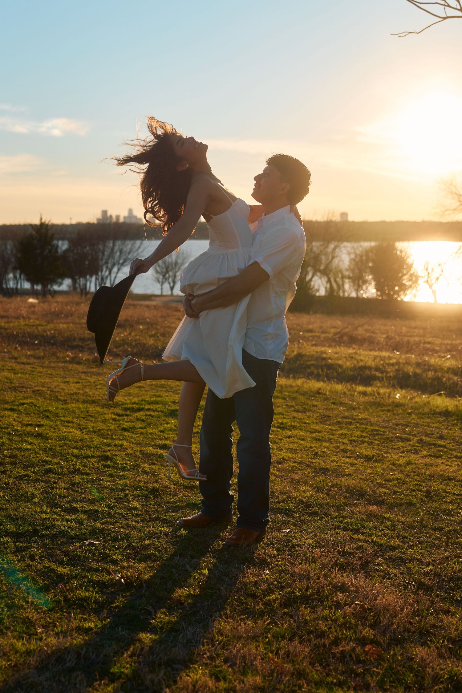 Couple embracing at golden hour sunset in a park, man lifting woman as her hair flows in the wind.
