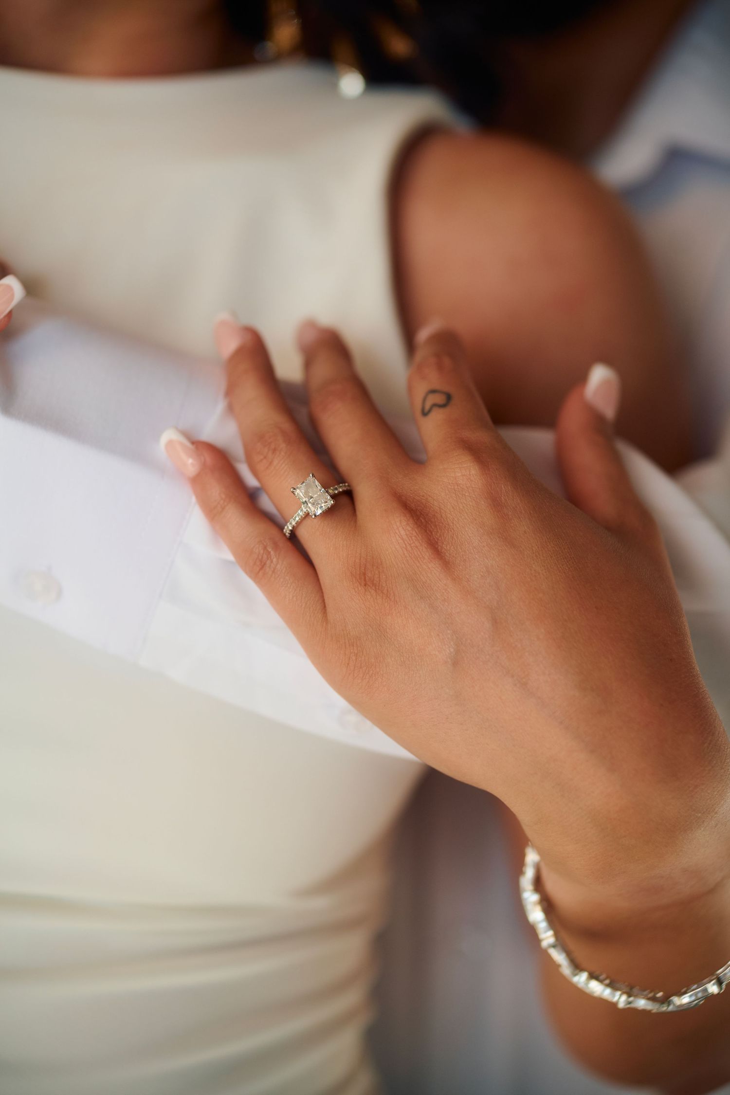 Close-up of a diamond engagement ring and bracelet on a woman's hand, with a small heart tattoo visible.