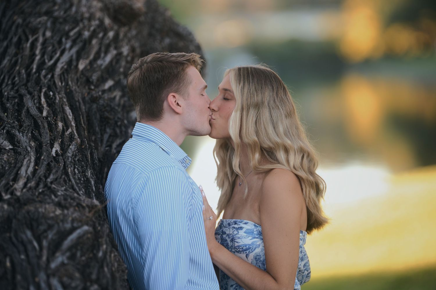 A romantic couple shares a tender moment against a large tree trunk at golden hour.
