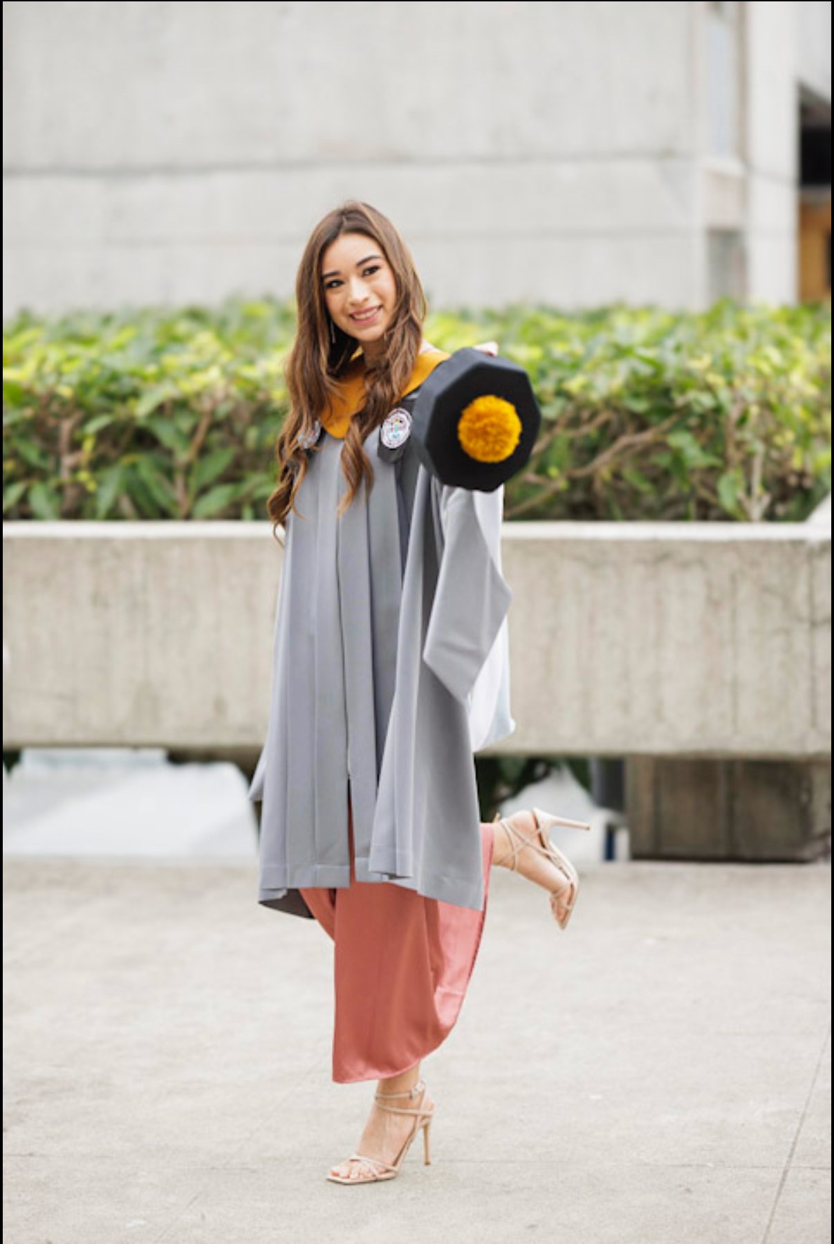 Graduate in grey academic regalia poses outdoors near concrete planters.