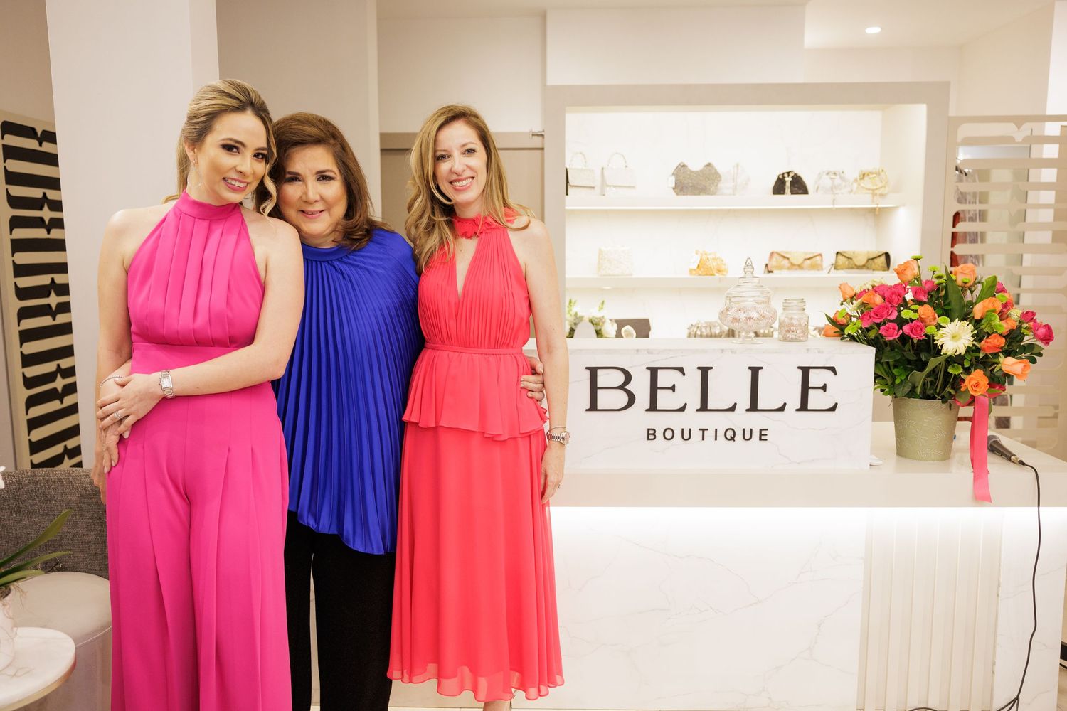 Women wearing vibrant pink and blue dresses stand together at Belle Boutique with retail displays.