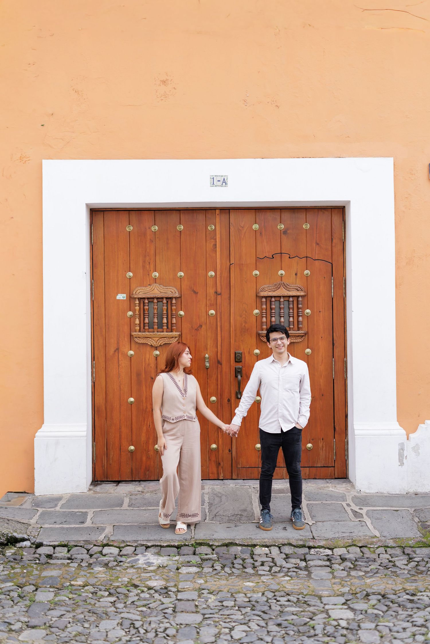 Stylish couple poses against peach wall with wooden doors in colonial setting.