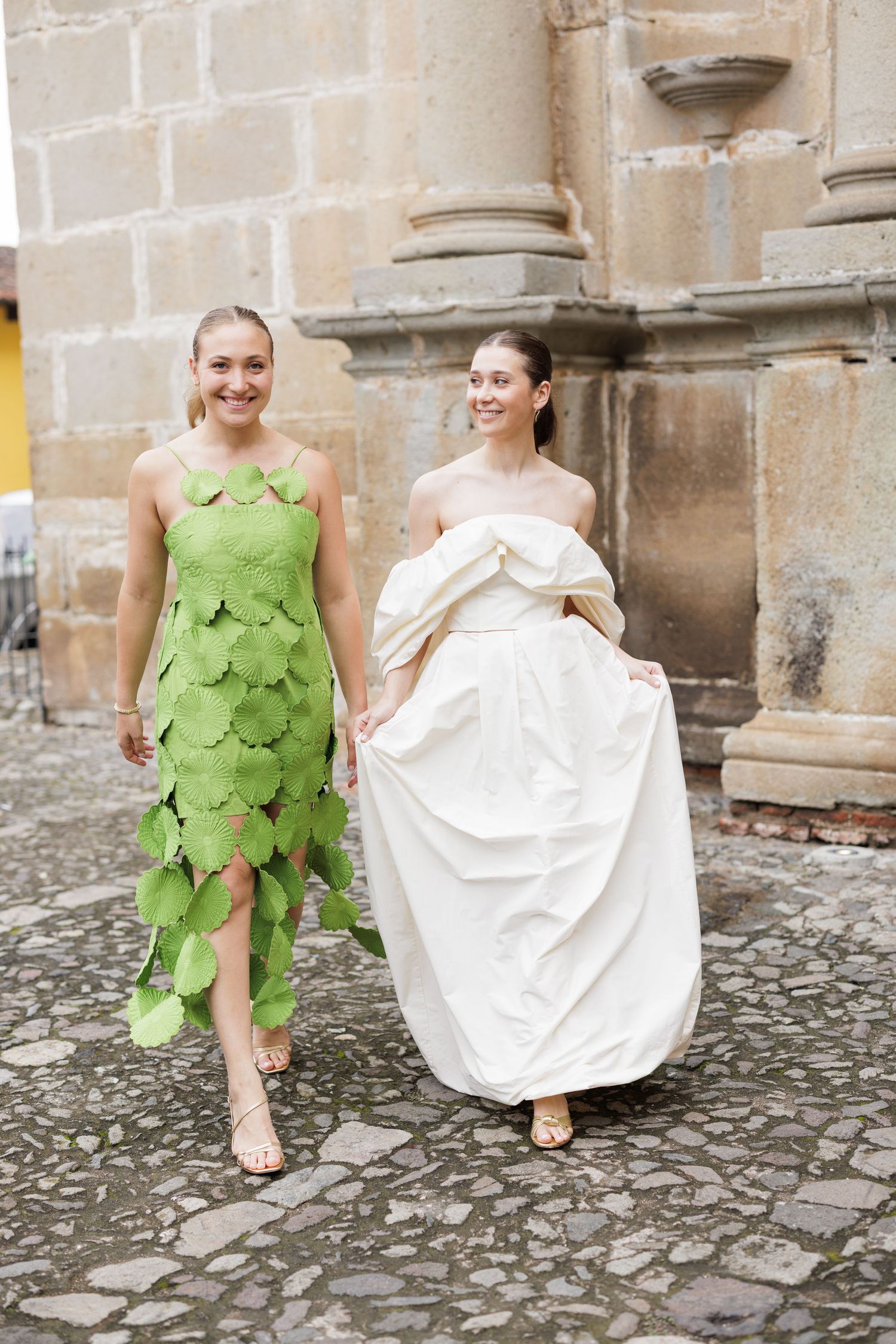 Two people in contrasting green and white dresses sitting on cobblestone steps outside a historic stone building.