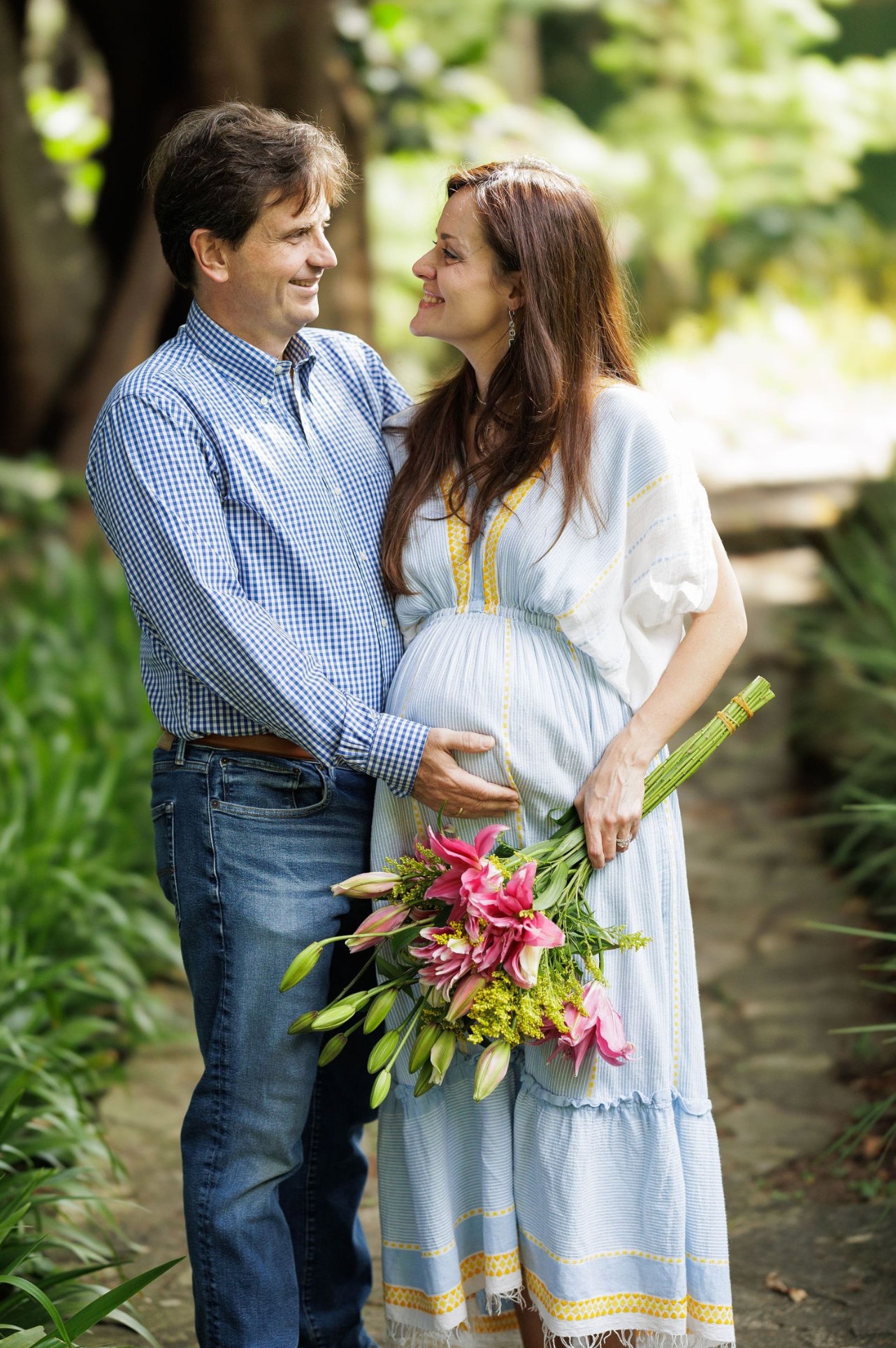 A couple in casual attire embraces outdoors while holding colorful flowers in a garden setting.