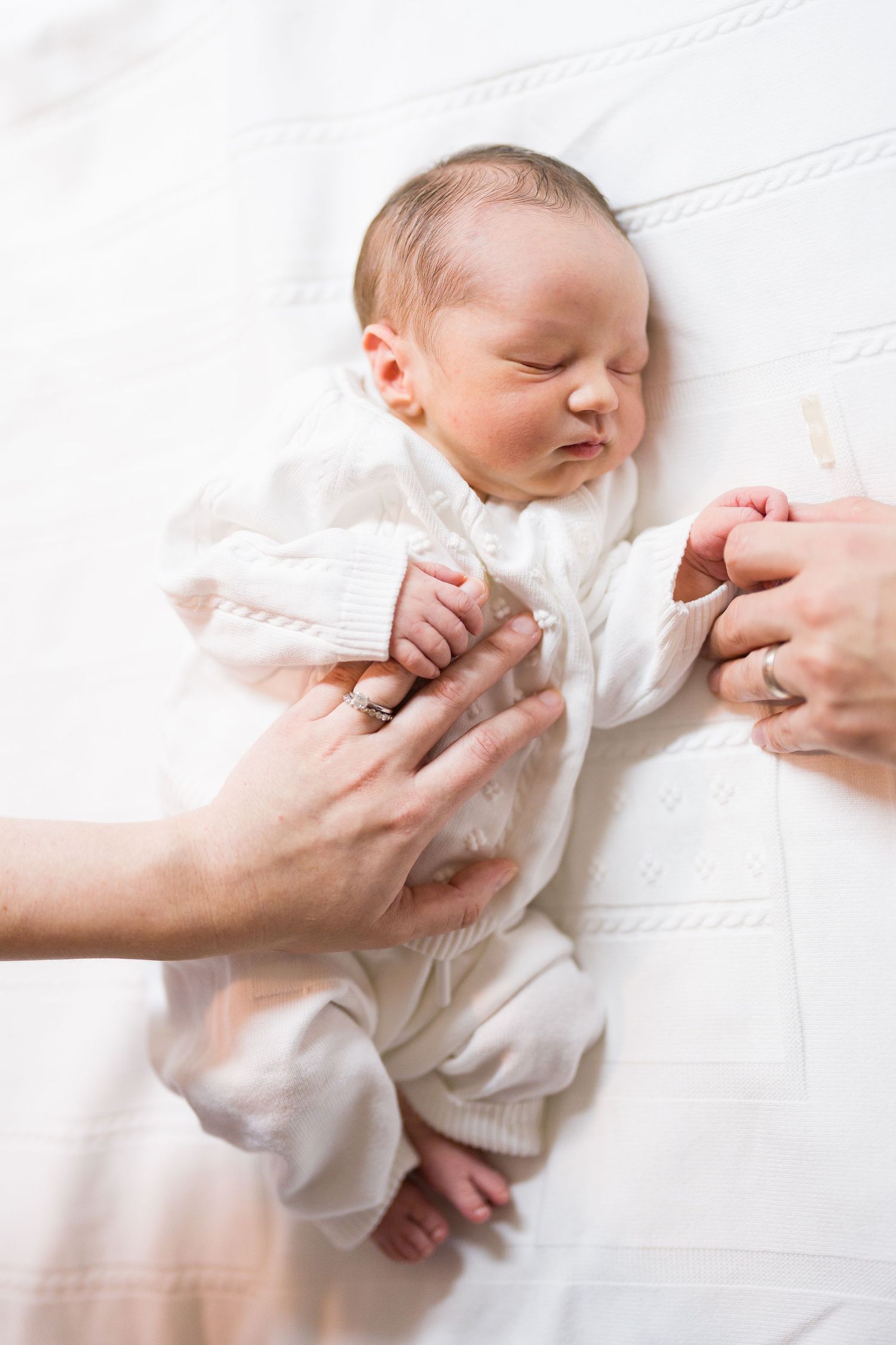 Caring hands tenderly hold sleeping infant wearing white sweater on white bedding.