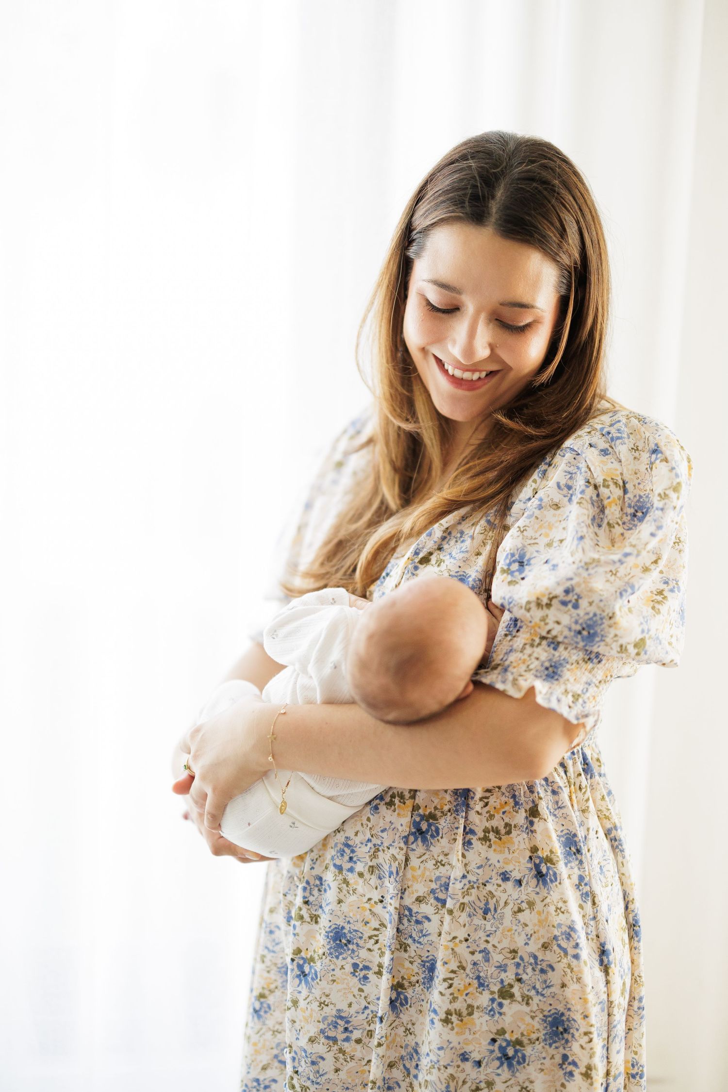 Person wearing floral dress holding and gazing at newborn baby against white backdrop.