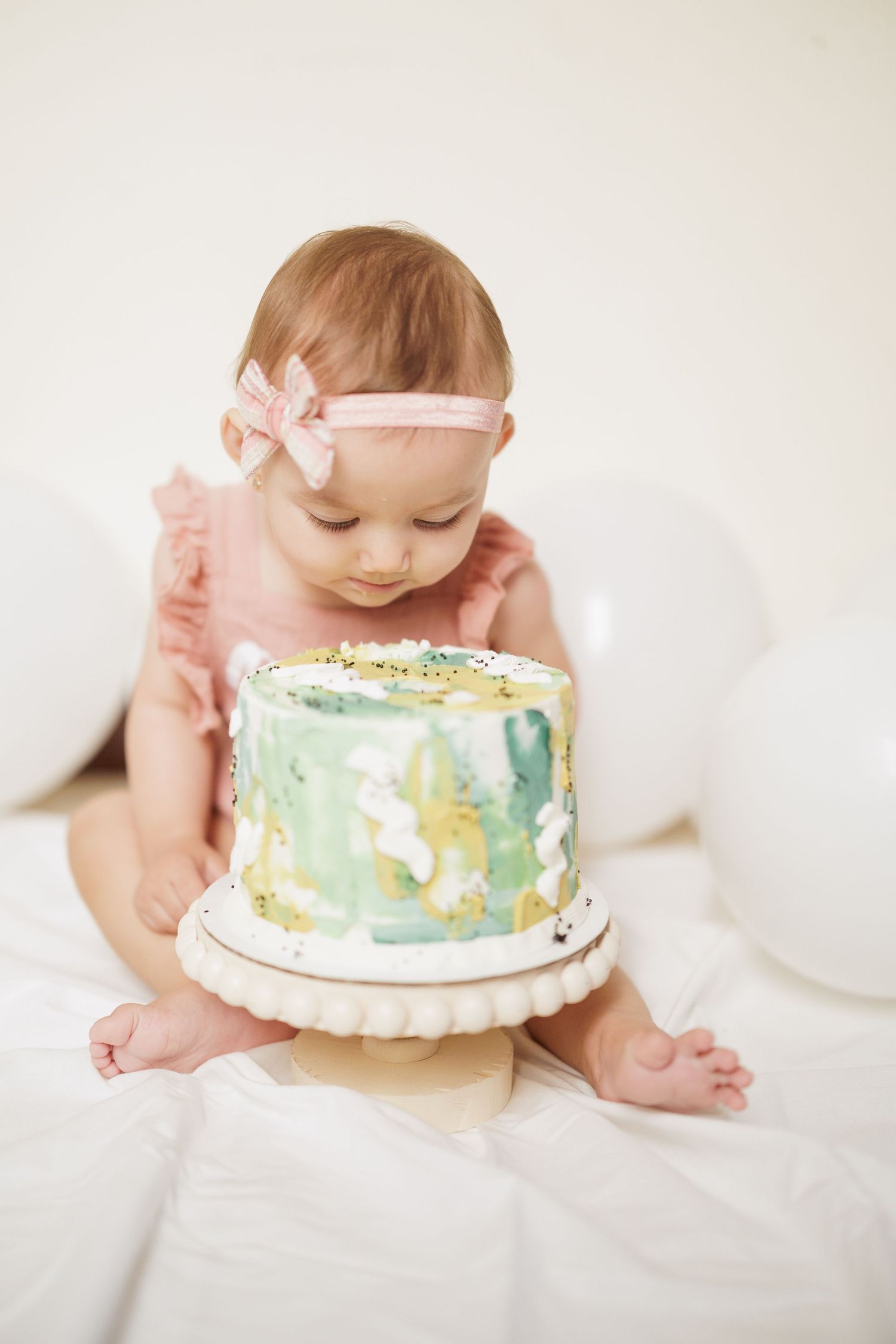 Baby exploring first birthday cake with pastel decorations on white background.