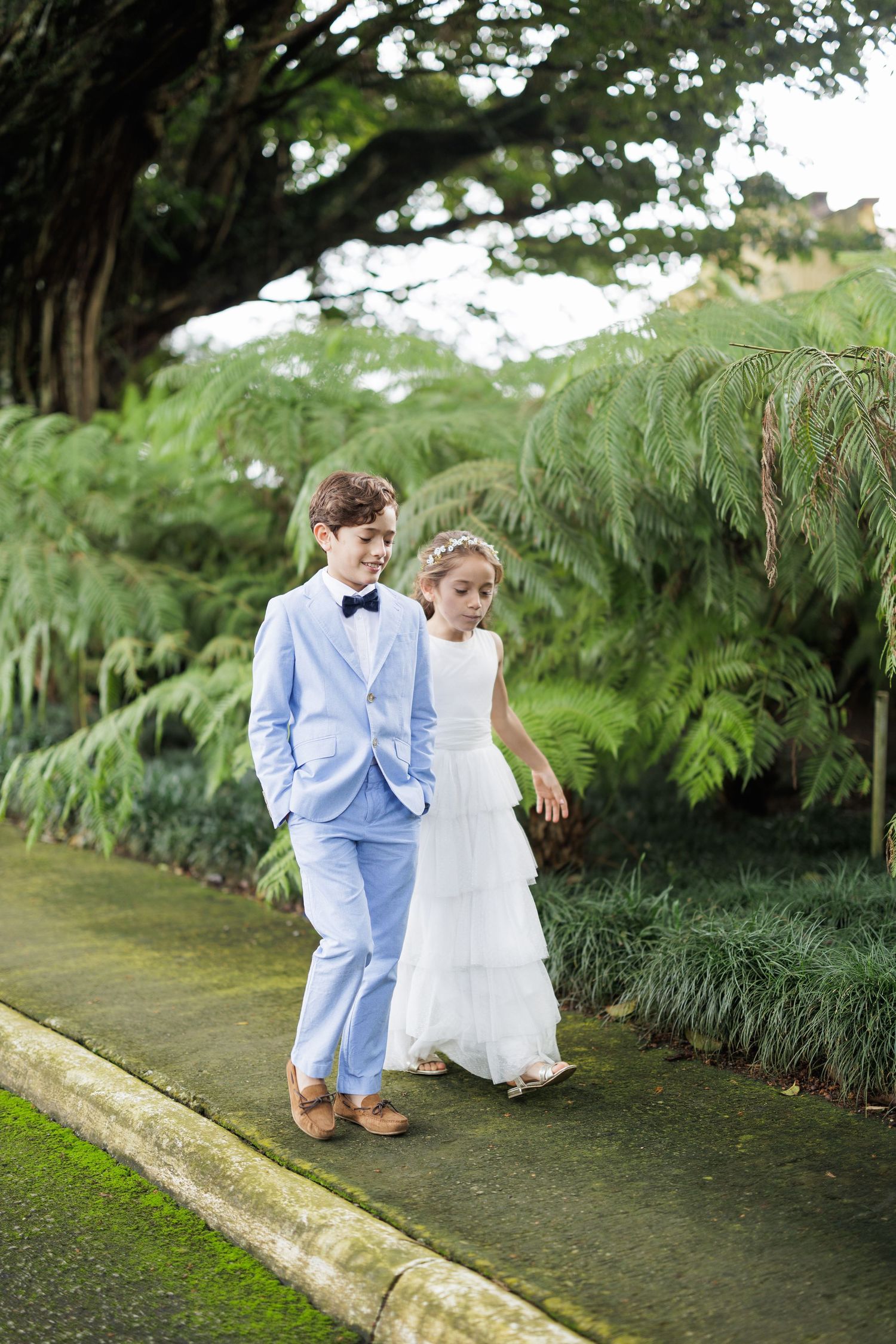Children in formal attire sit together on stone path surrounded by lush greenery.