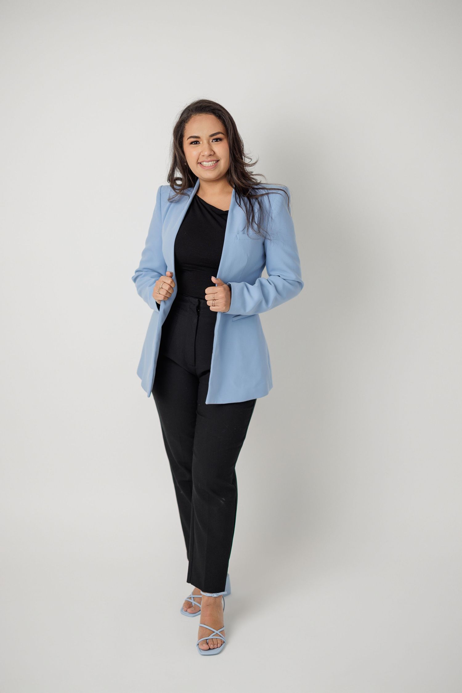 Fashion model poses in black dress and light blue blazer against neutral studio backdrop.