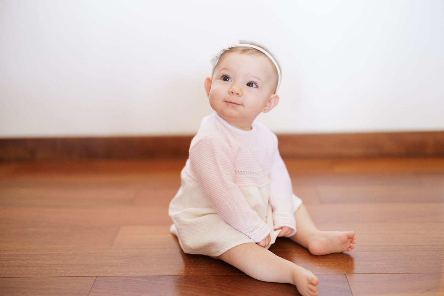 Baby in cream-colored outfit sitting on hardwood floor.