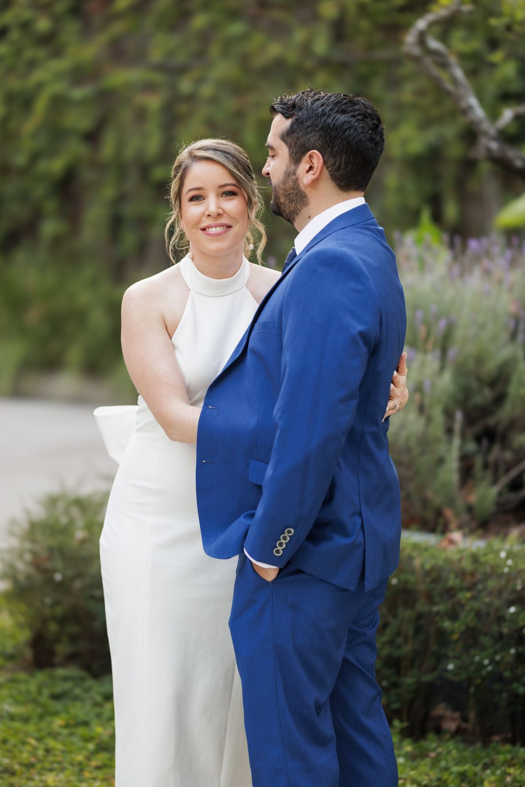 Newlyweds in white dress and navy suit embrace in an outdoor garden setting.