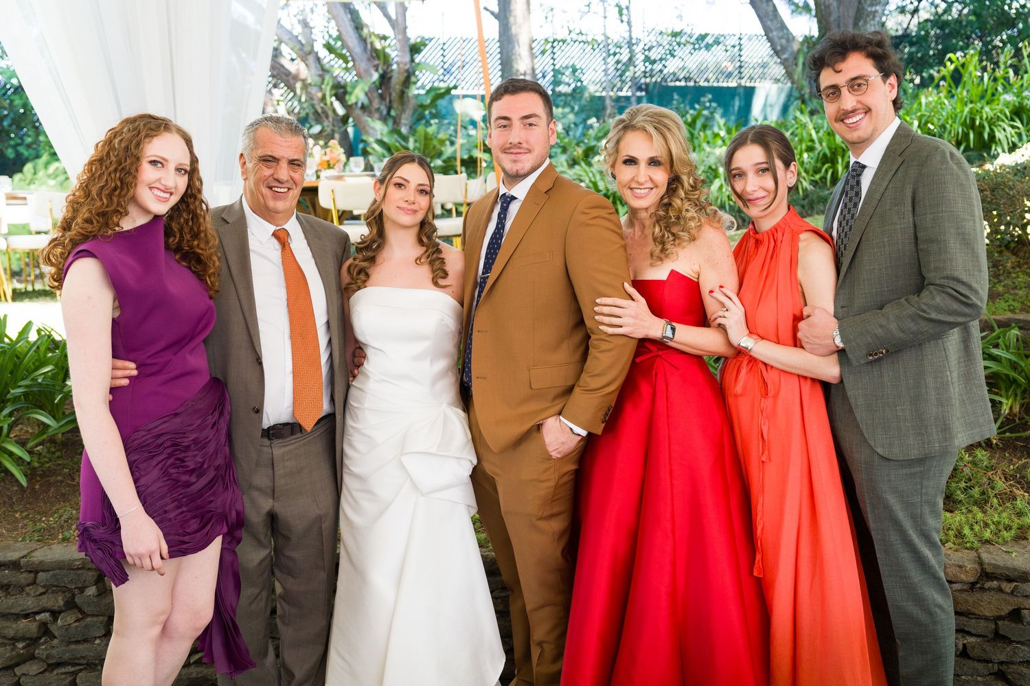 Wedding party wearing vibrant red and coral dresses pose by stone wall outdoors.