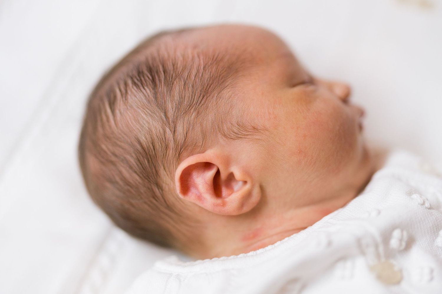 Sleeping newborn baby wearing white clothing rests peacefully on white bedding.