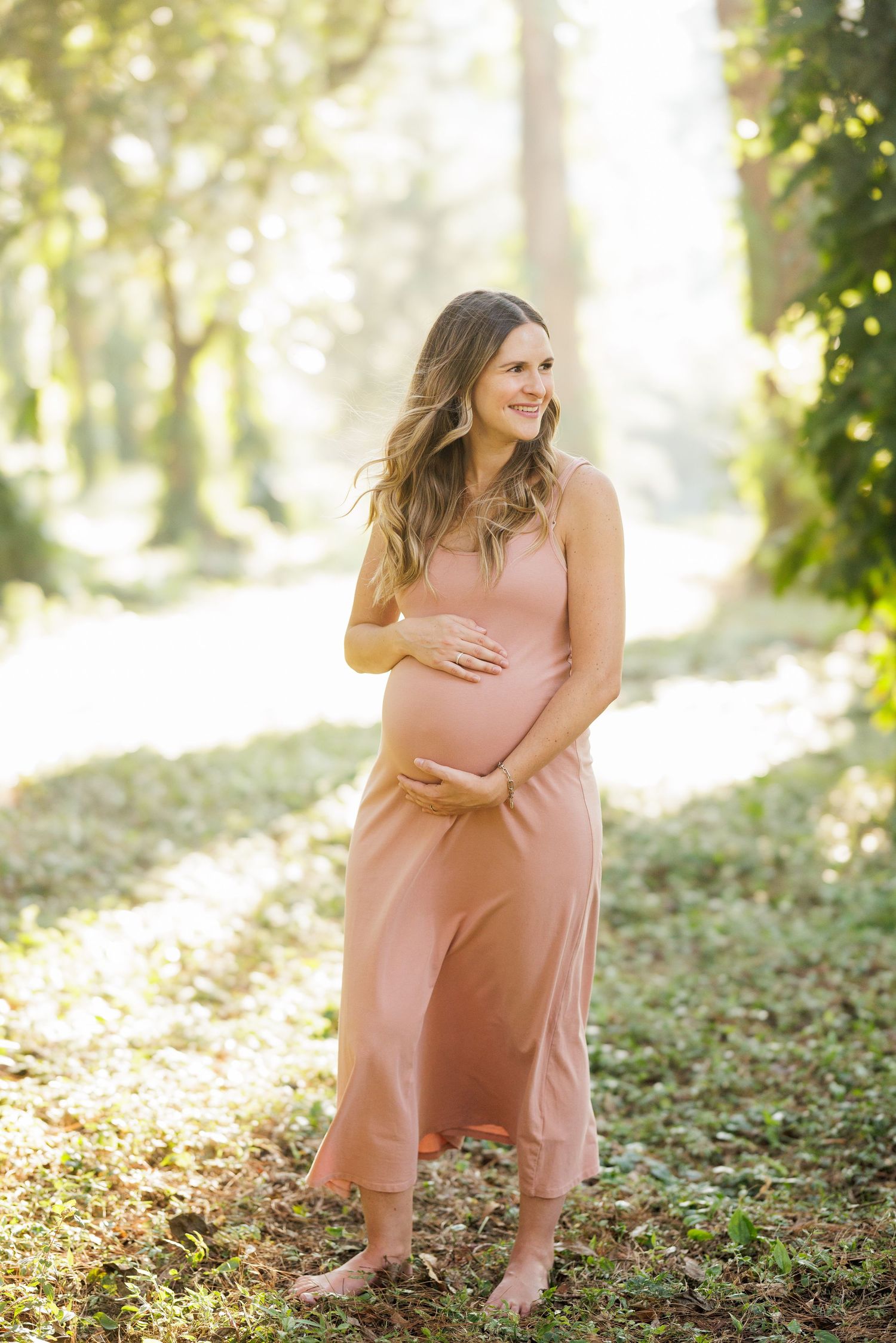 Maternity portrait session in a sunlit forest with woman wearing a pink flowing dress.