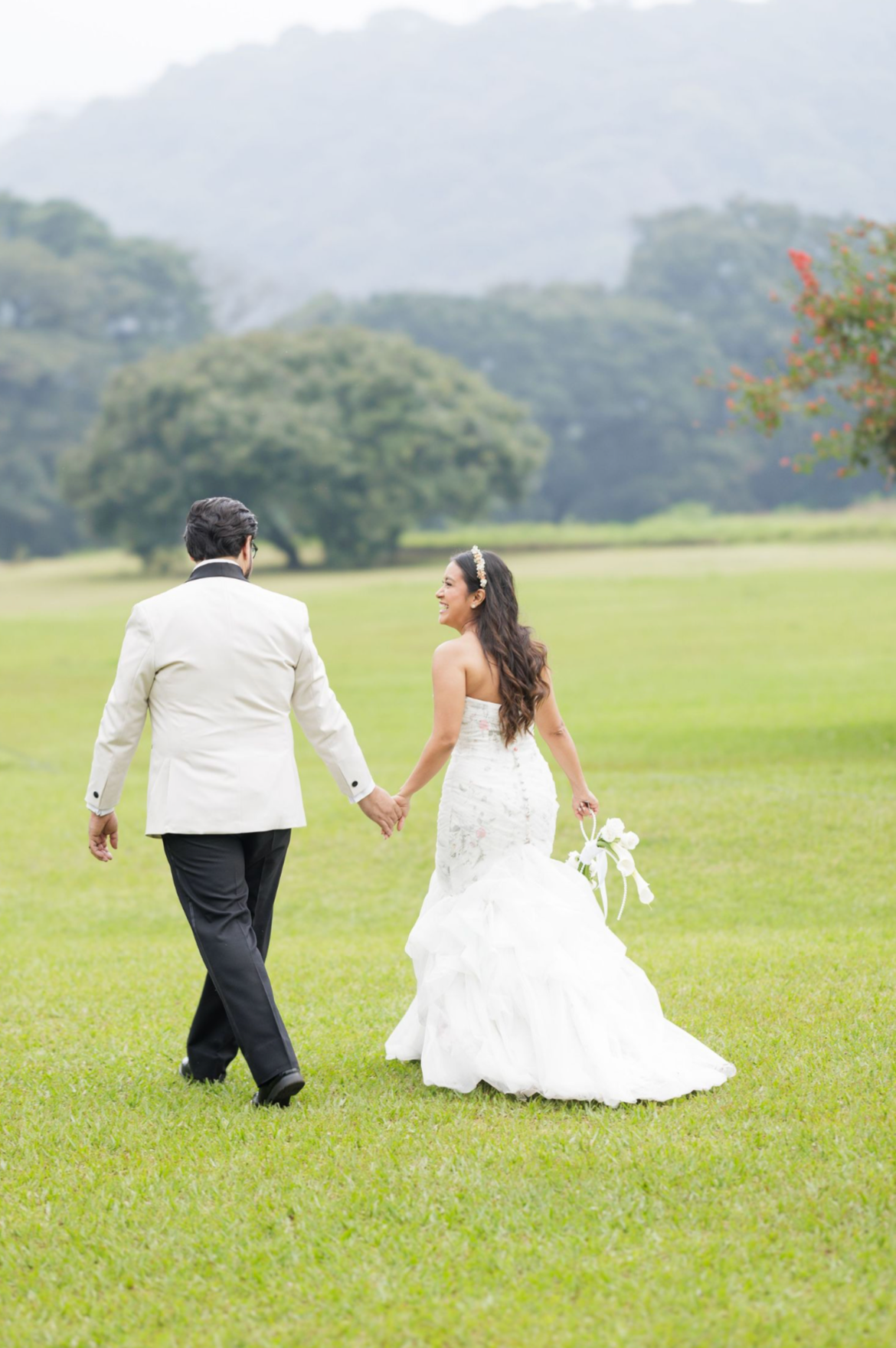 Newlyweds walk hand in hand across a green field with mountains in the background.
