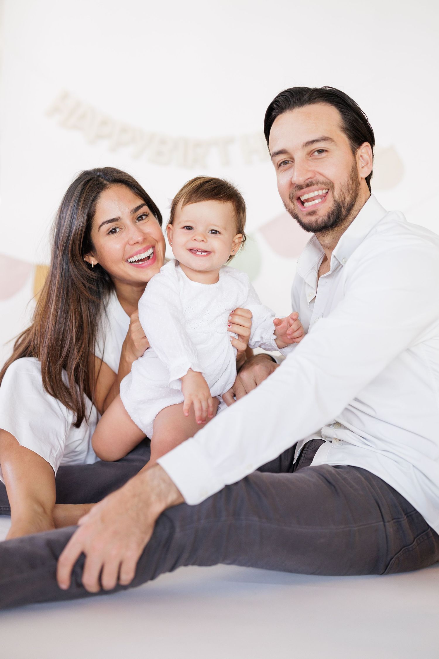 Family lounging together on white bedding in bright natural light.