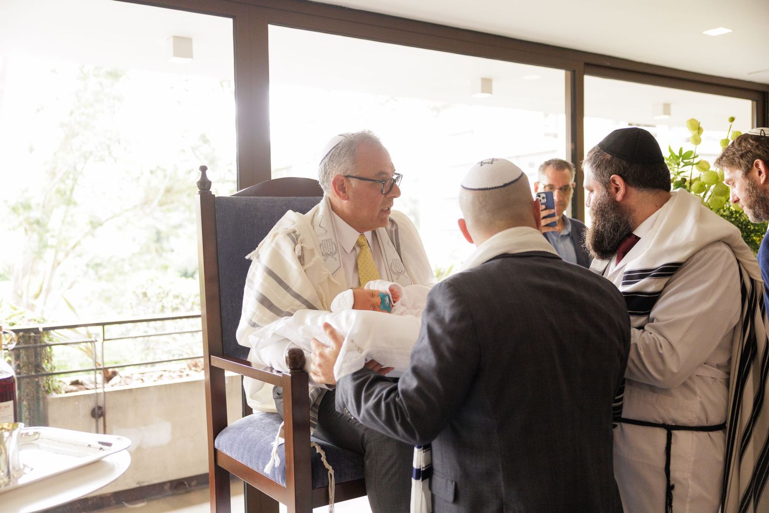 A group gathers around a table during a religious ceremony in a bright sunlit room.