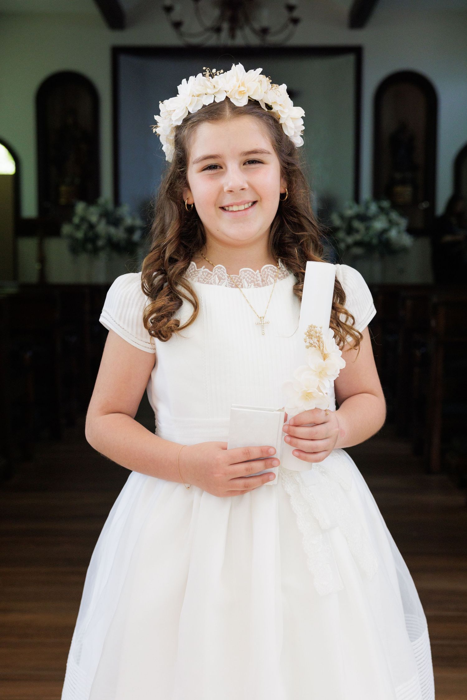 A flower girl in a white dress and floral crown stands indoors at a formal event.