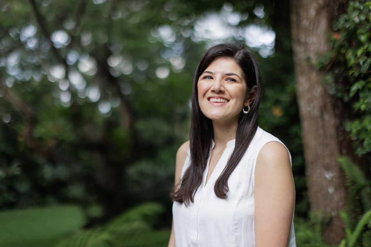 Woman in white sleeveless top smiling outdoors with natural greenery background.