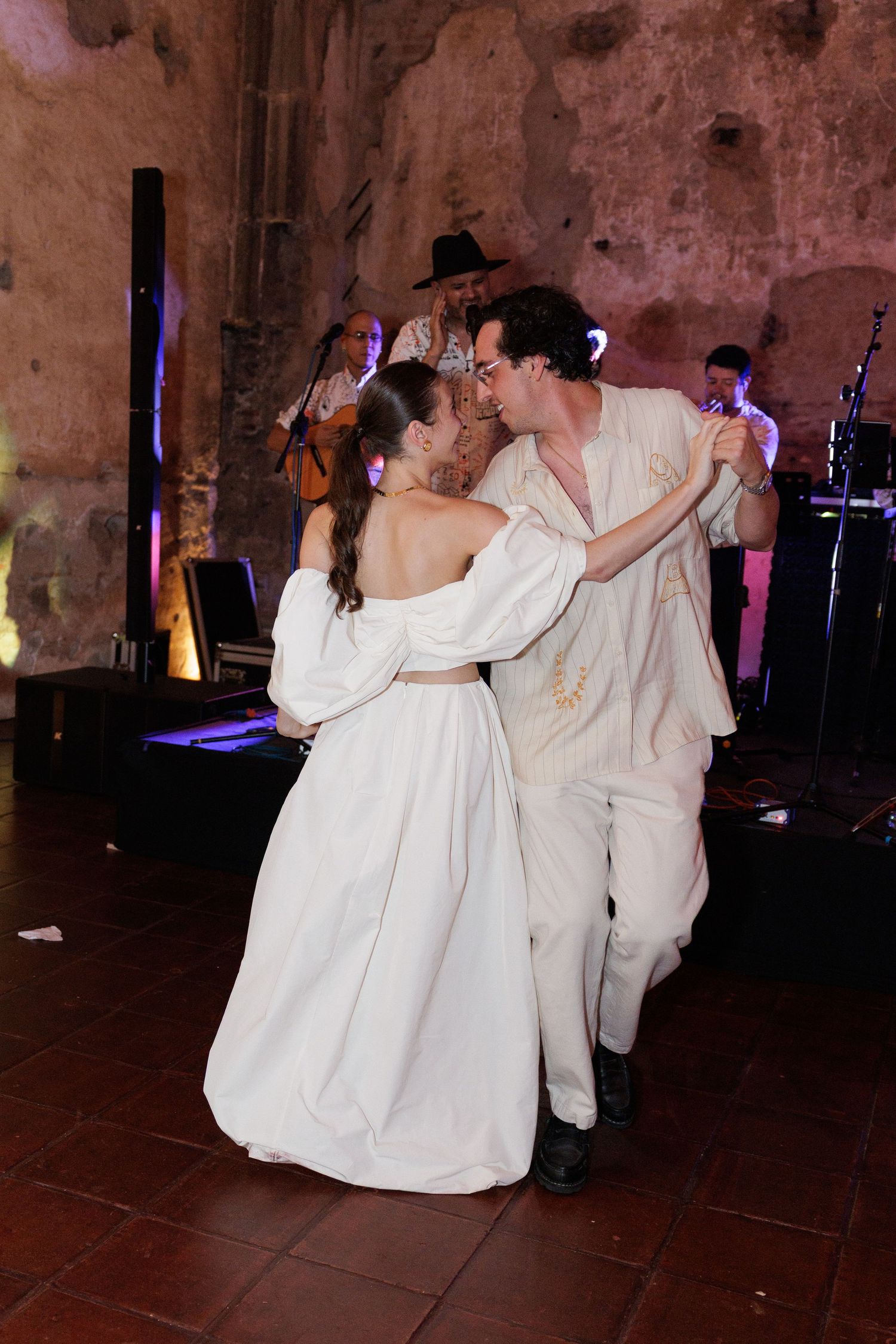 Dancing figures in white attire enjoying a celebration in a dimly lit historic venue.