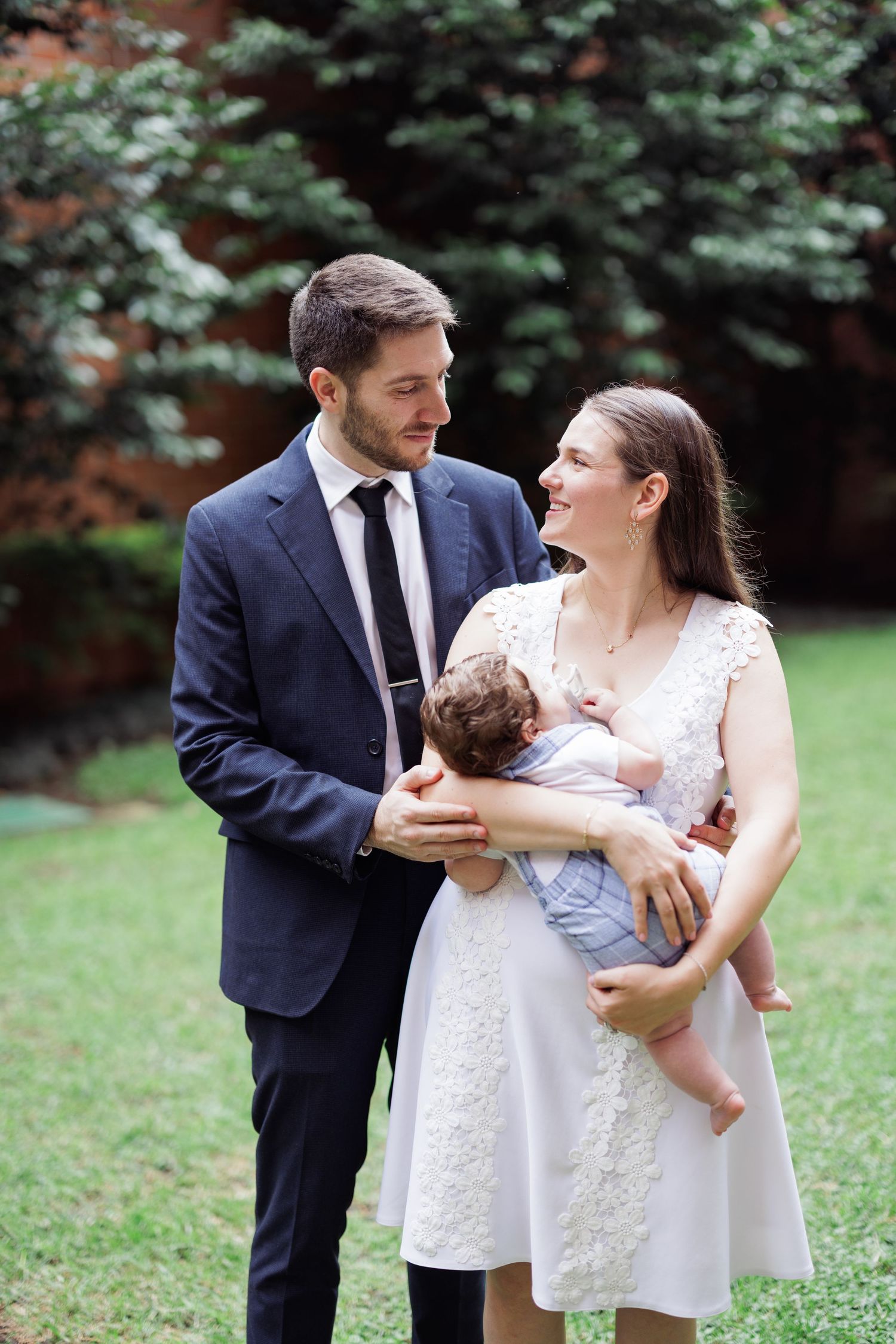 A couple in formal attire shares sweet moments with their baby in a garden setting.