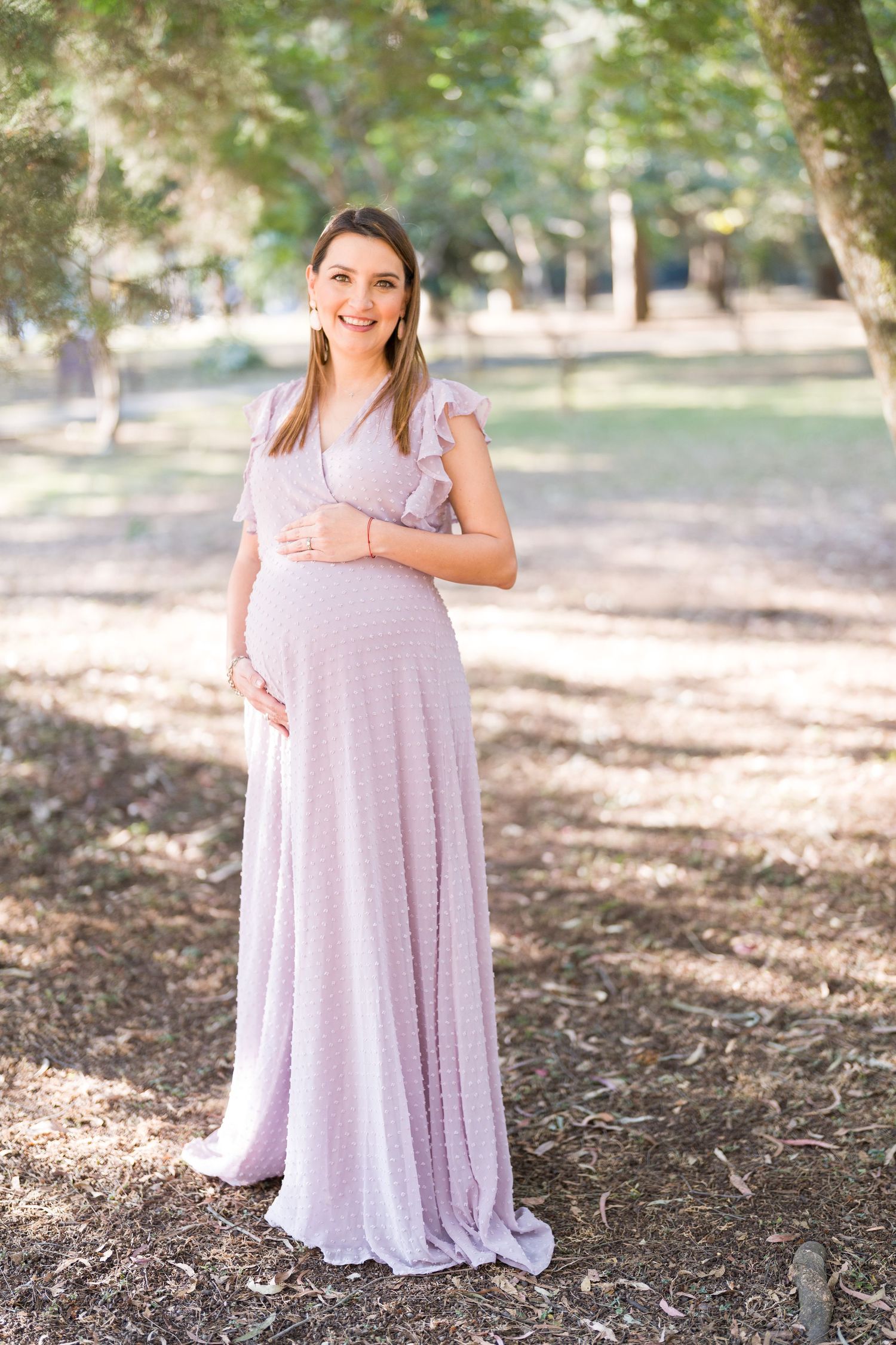 Woman in a flowing lavender dress poses in a wooded setting with dappled sunlight.