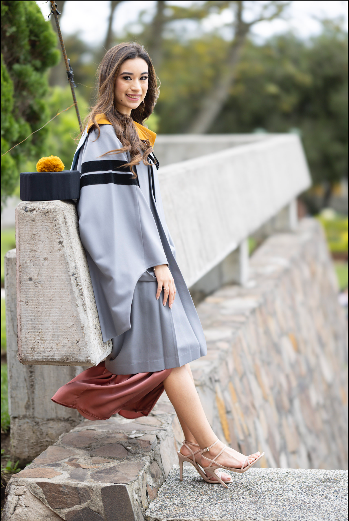 Graduate in grey graduation gown poses on stone wall during photo session.