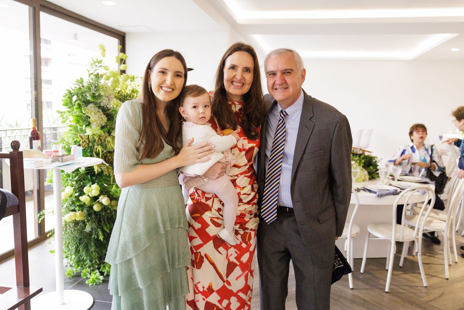 Three people pose together at an elegant indoor celebration with floral decorations and white tables in the background.