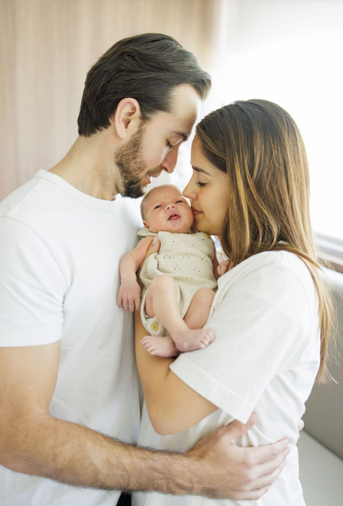 A tender family moment with parents embracing their baby in white outfits against a bright indoor backdrop.