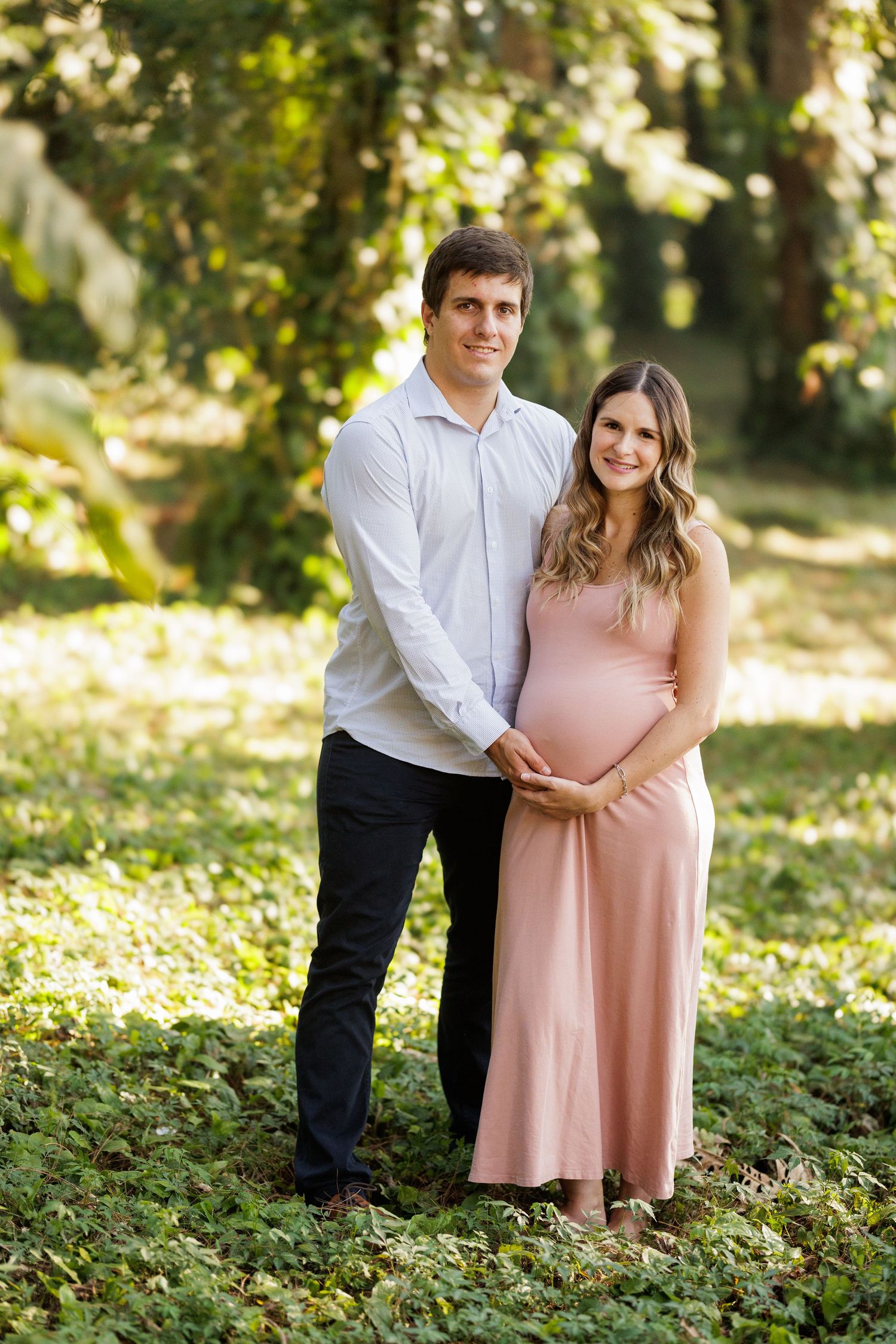 A couple poses together outdoors in a sunlit wooded area wearing pink and white clothing.