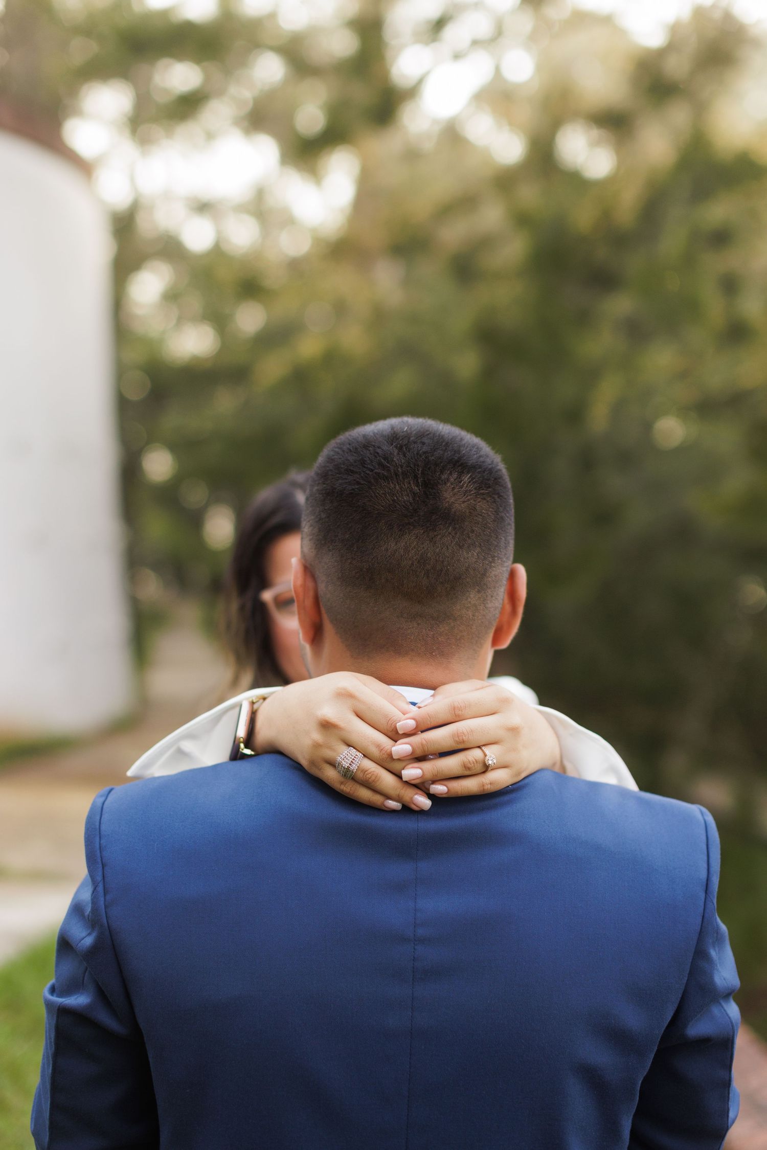 Close-up of hands embracing on a navy suit jacket from behind in an outdoor setting.