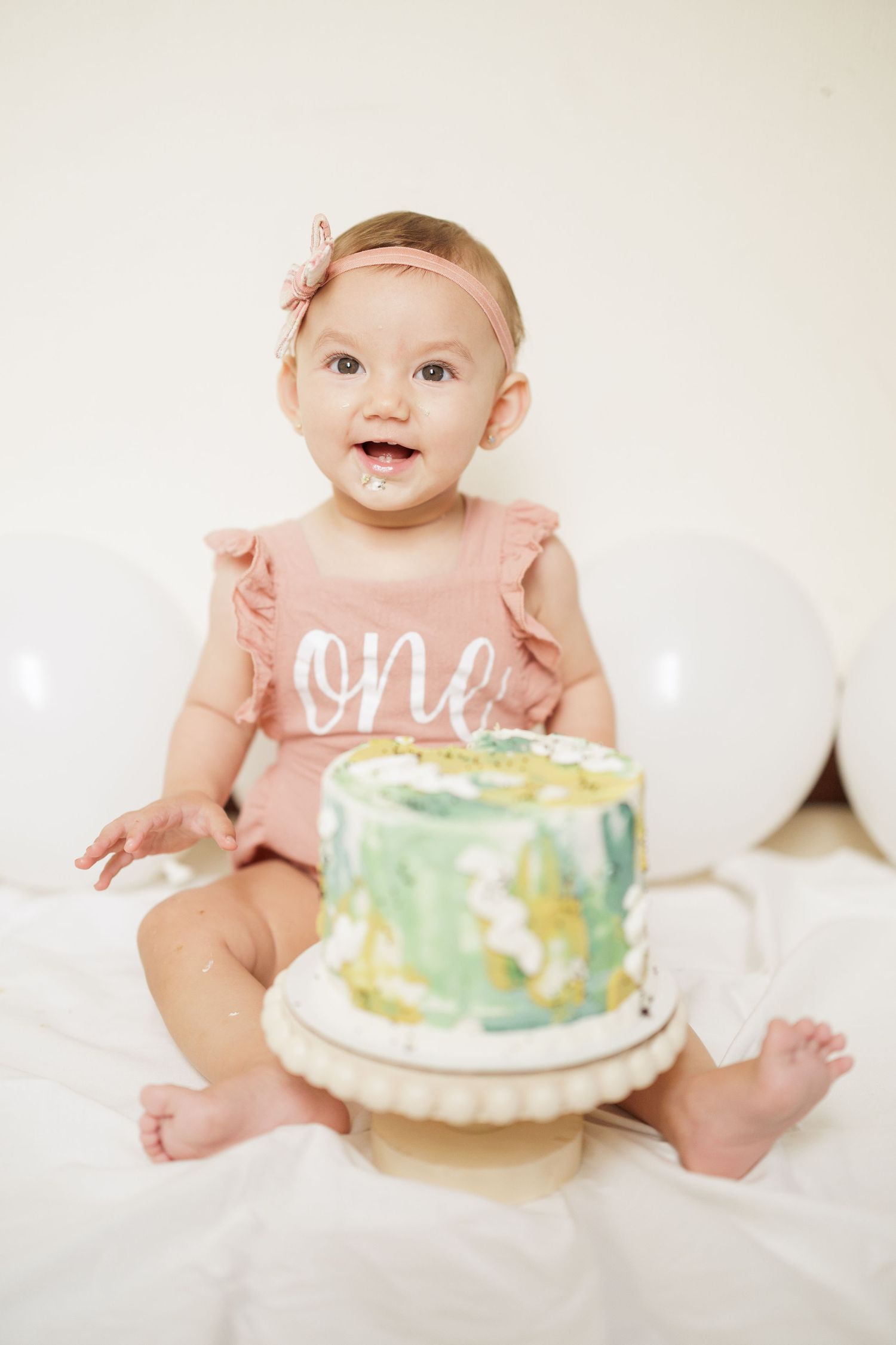 Baby in pink outfit sitting with decorated pastel birthday cake and white balloons.