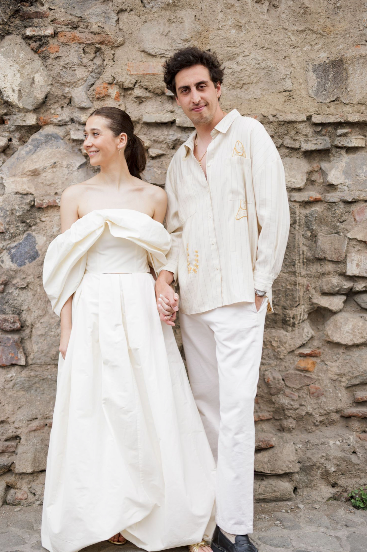 Couple in all-white outfits poses against stone wall during wedding photoshoot.
