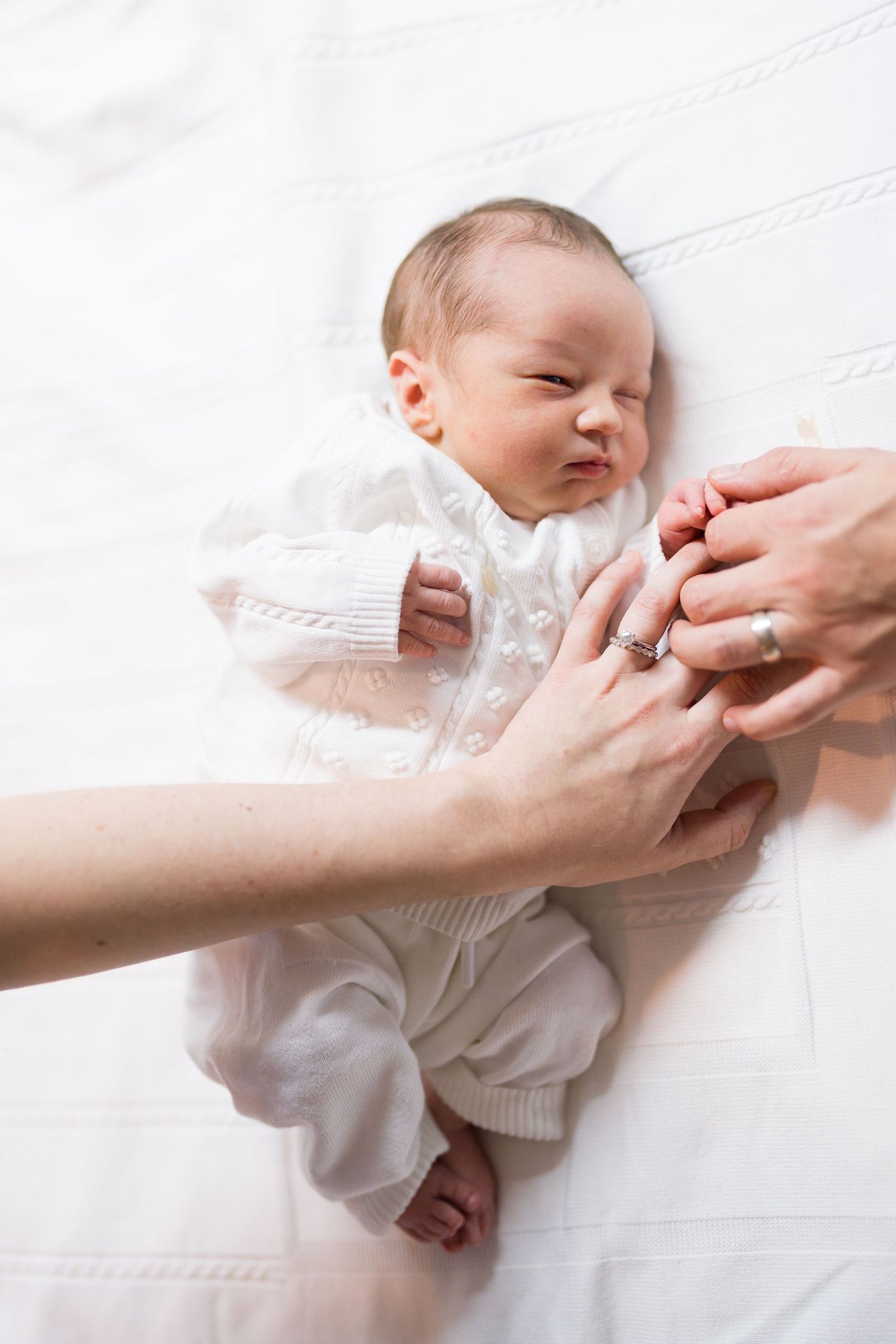 Gentle hands cradle a sleeping newborn baby dressed in white on soft bedding.