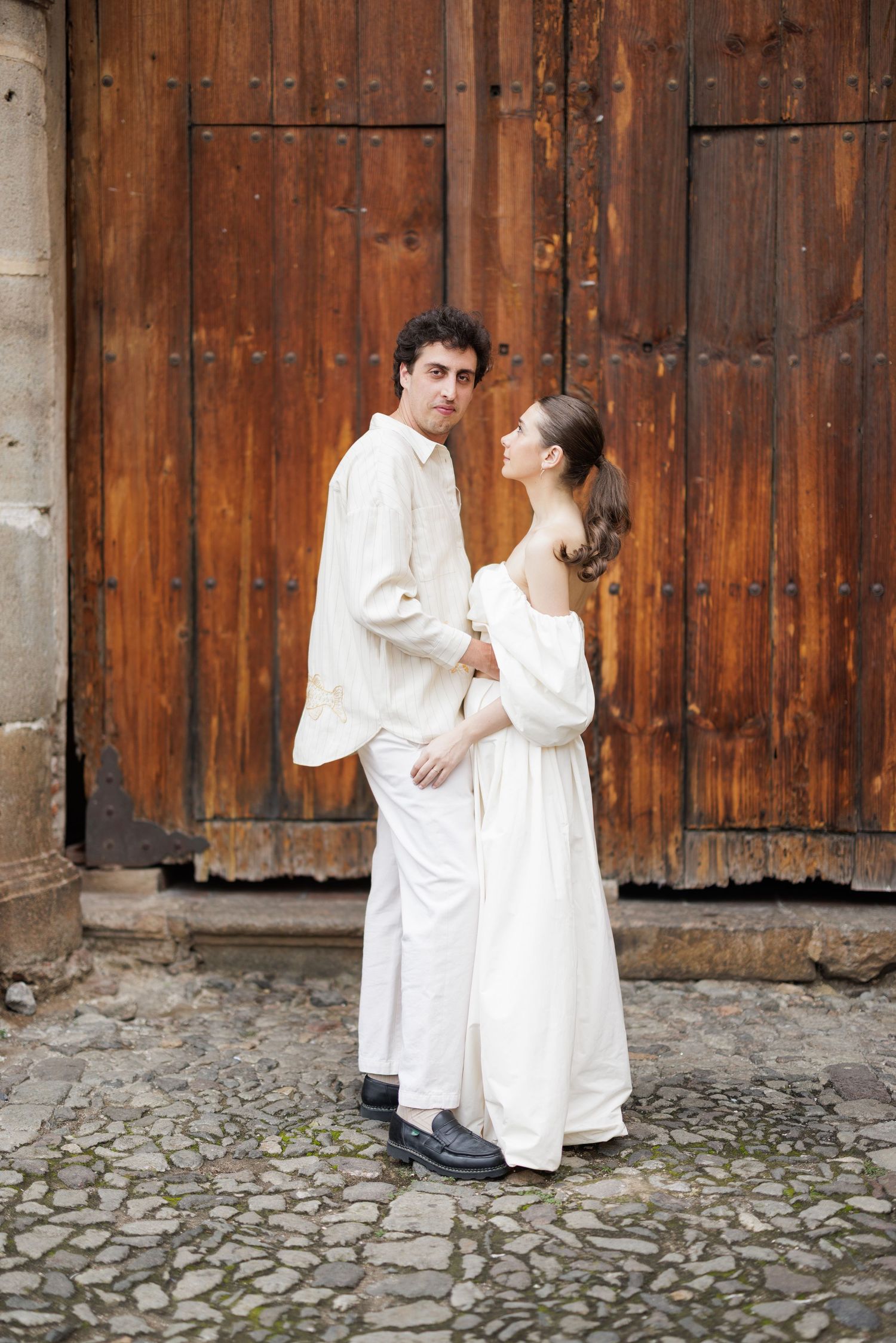 Couple in white outfits sharing playful moments against a rustic wooden door backdrop.