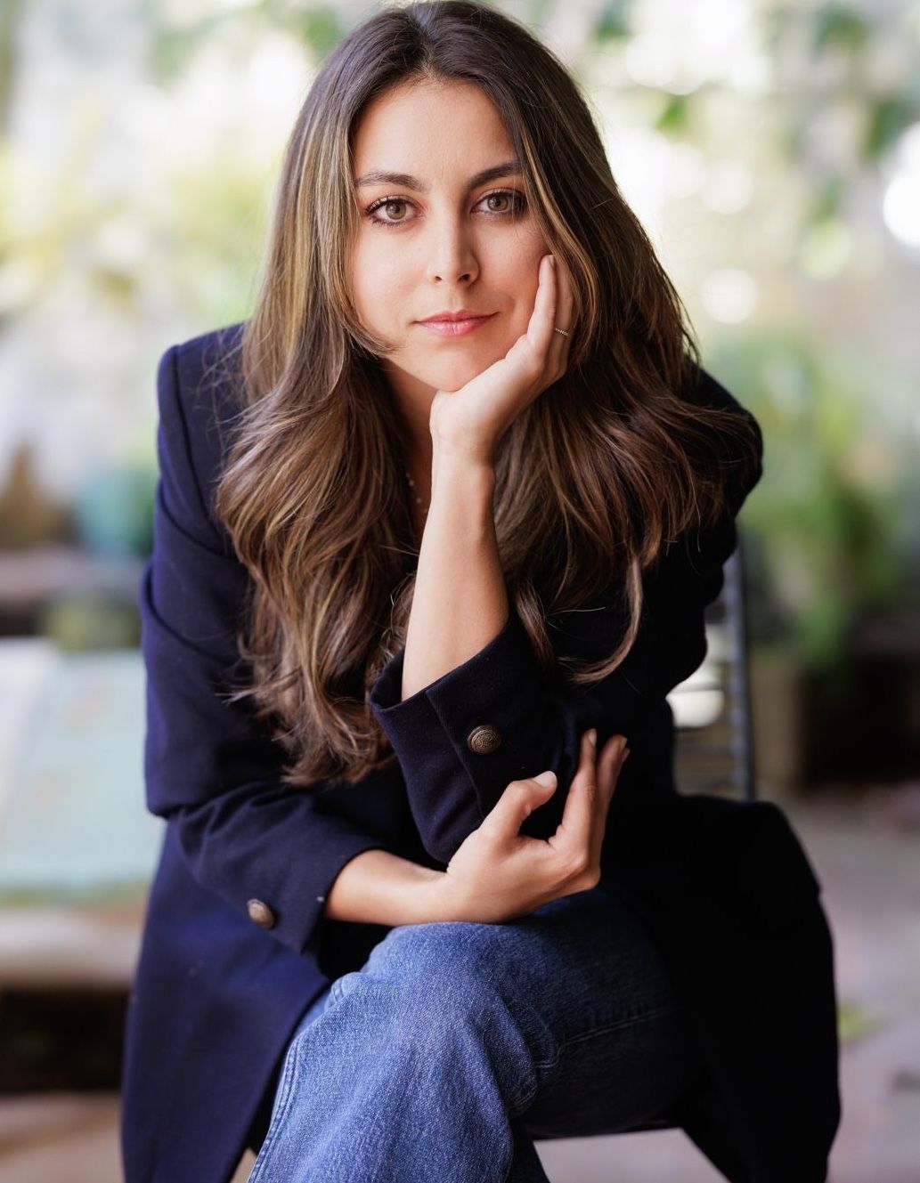 Woman in navy blazer poses thoughtfully with hand on face in outdoor setting.