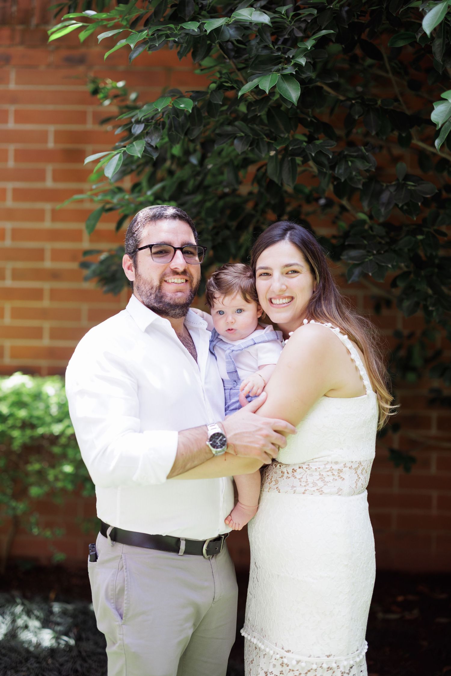 A family shares tender moments outdoors against a brick wall backdrop in matching white outfits.