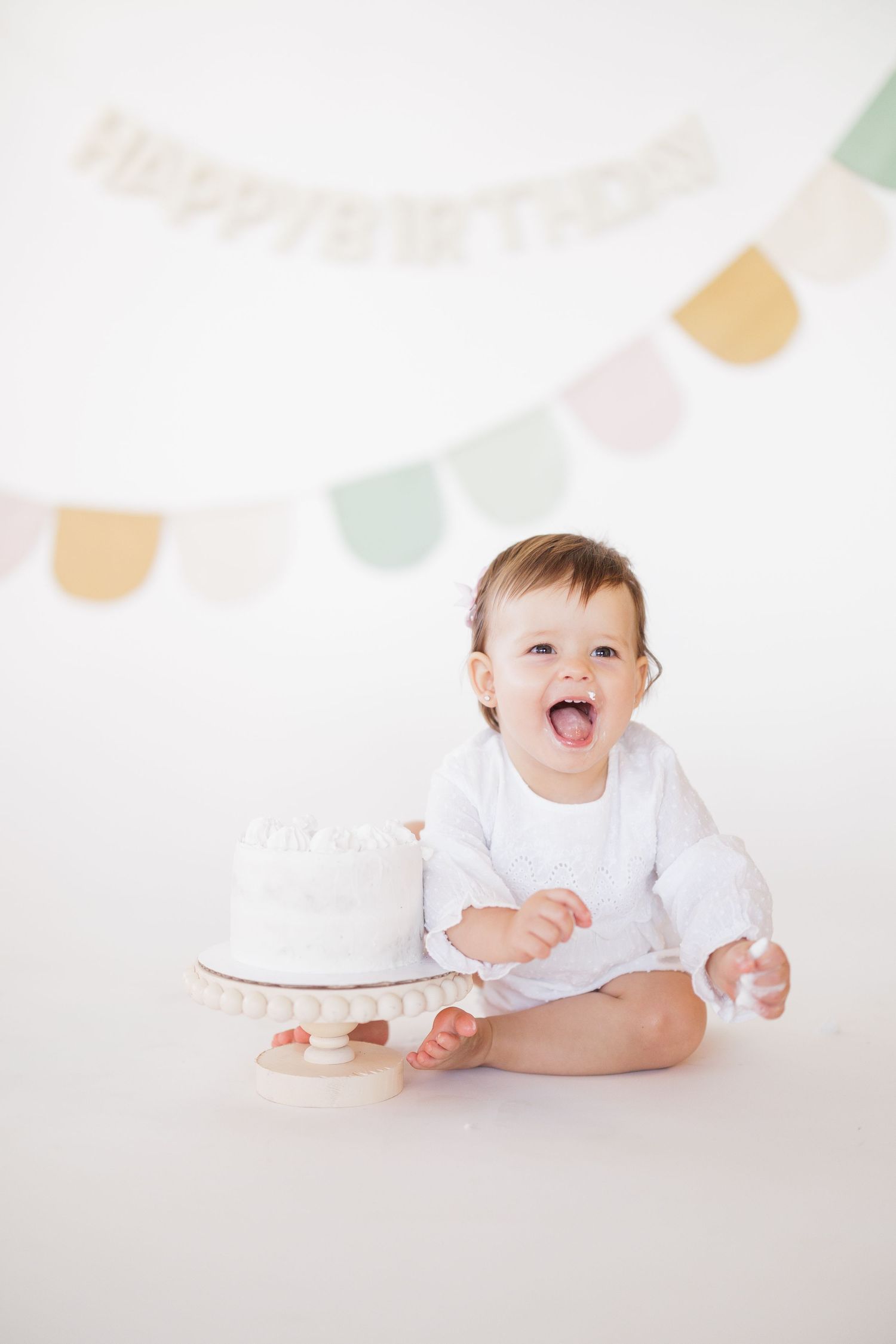 Baby in white outfit playing with cake during first birthday photoshoot with pastel bunting.