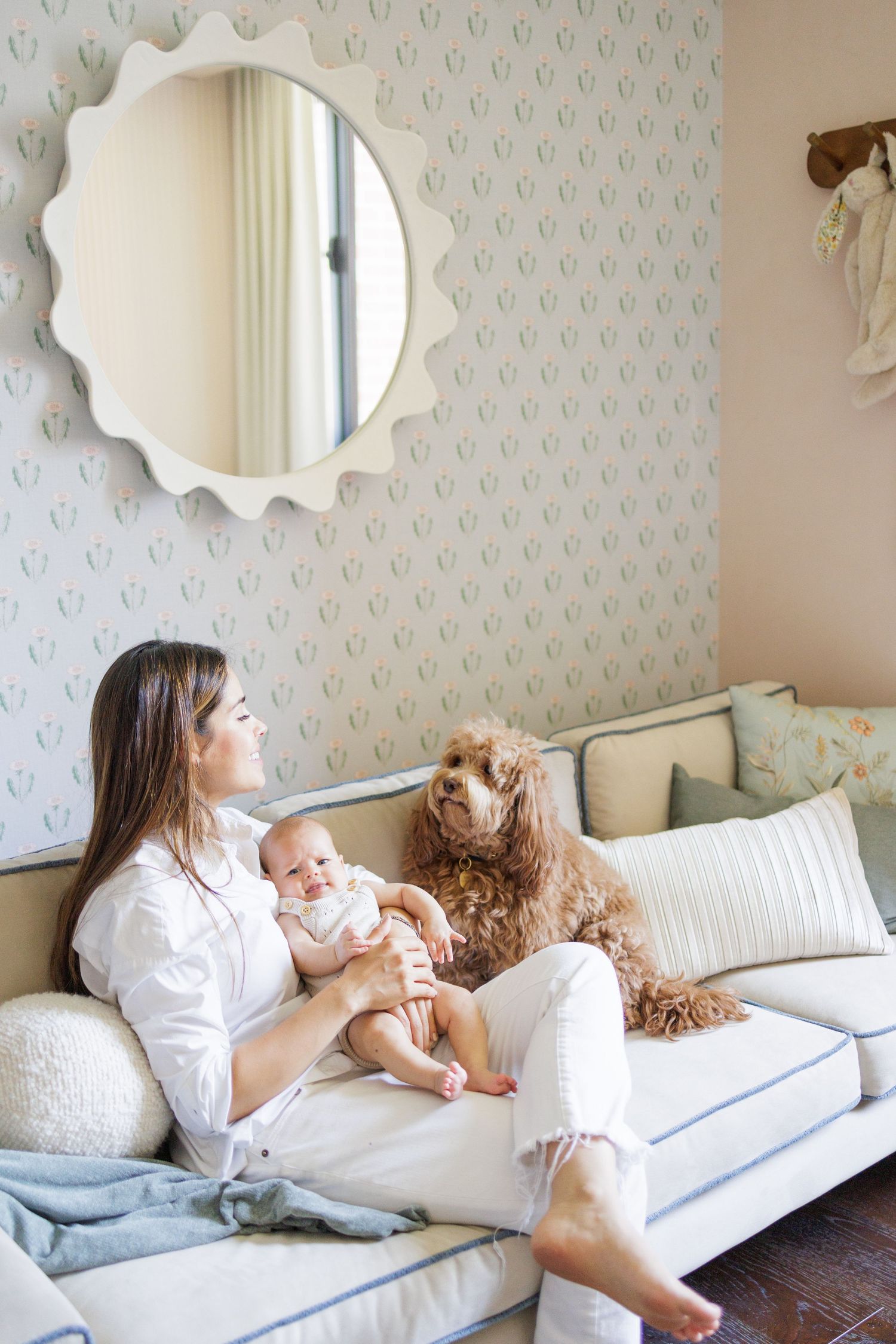 Mother and baby relax on white couch with teddy bear in cozy room with patterned wallpaper.