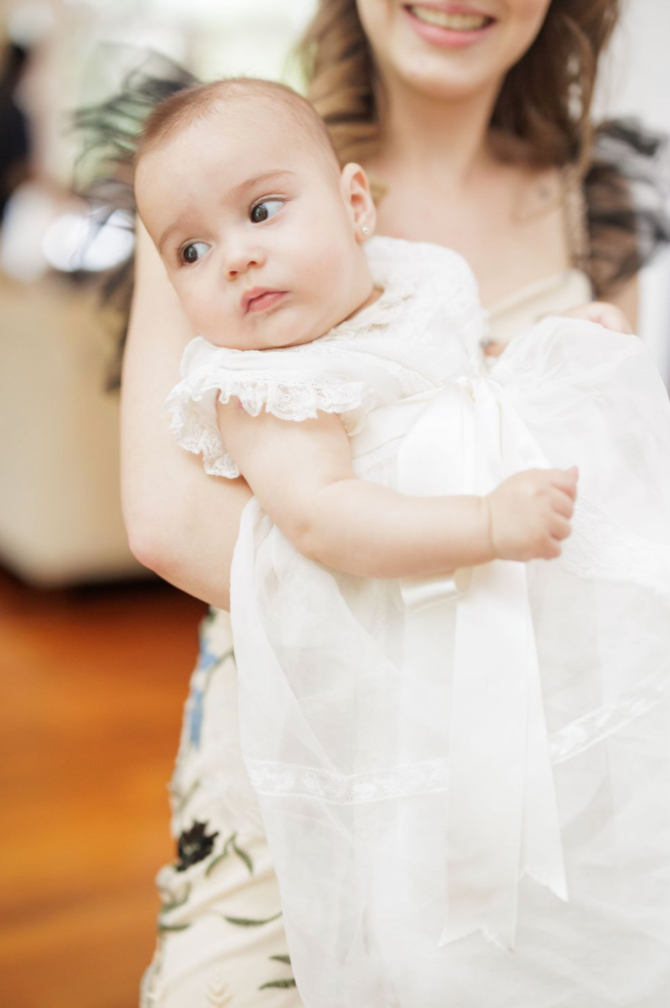 Baby wearing white christening gown looks at camera with curious expression.