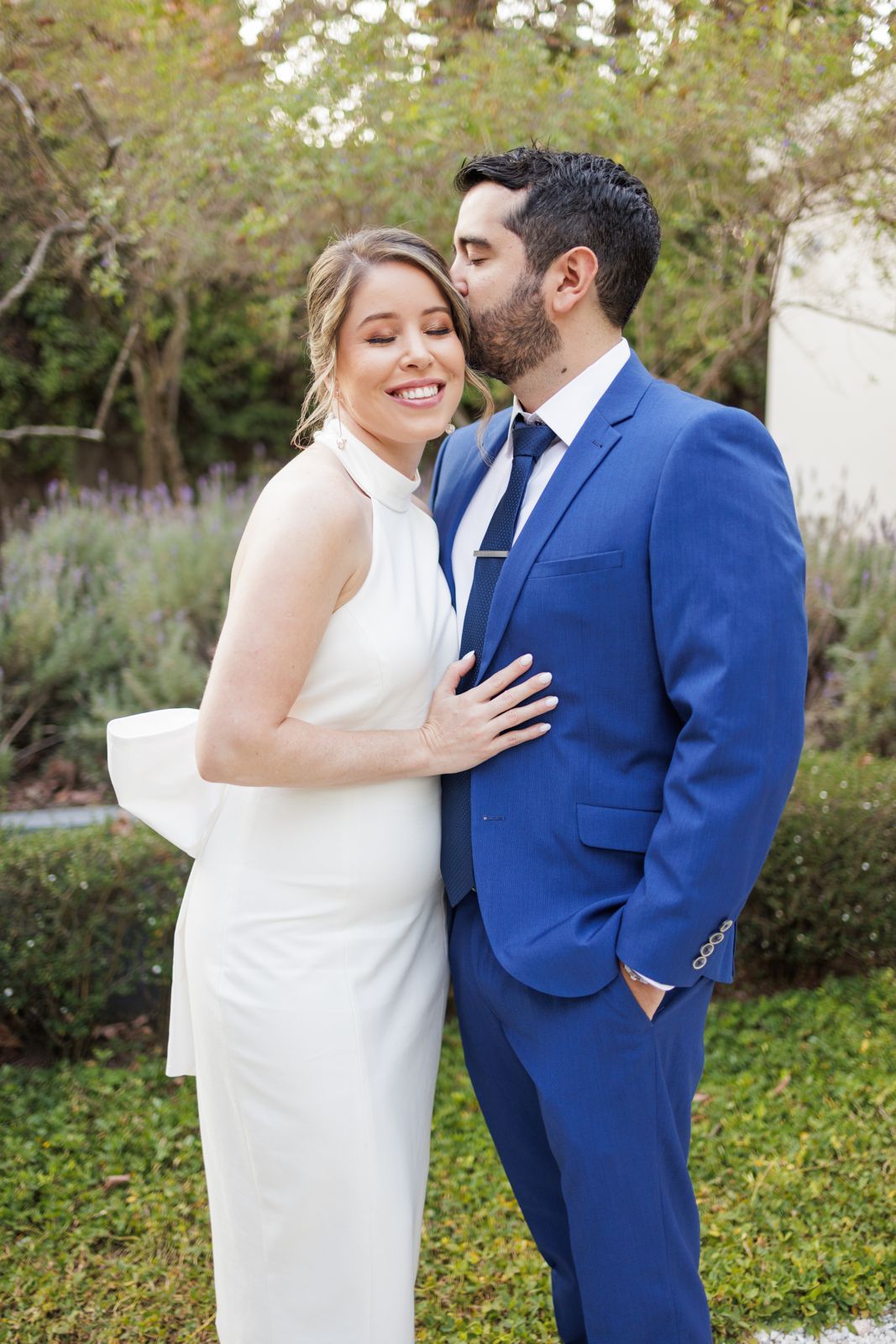 Couple in elegant white dress and blue suit embrace outdoors.