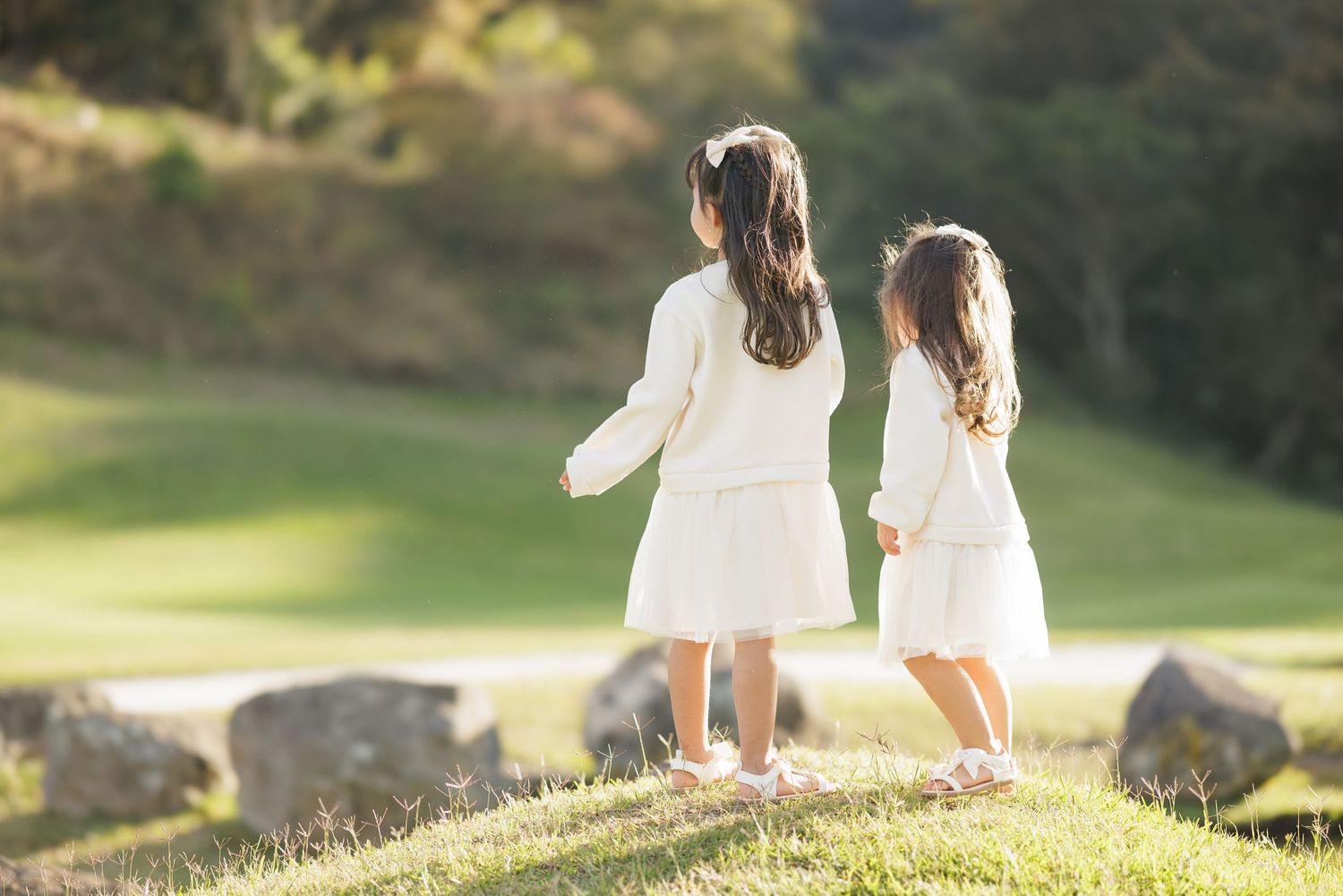 Mother and daughter in matching white dresses hold hands on grassy hillside.