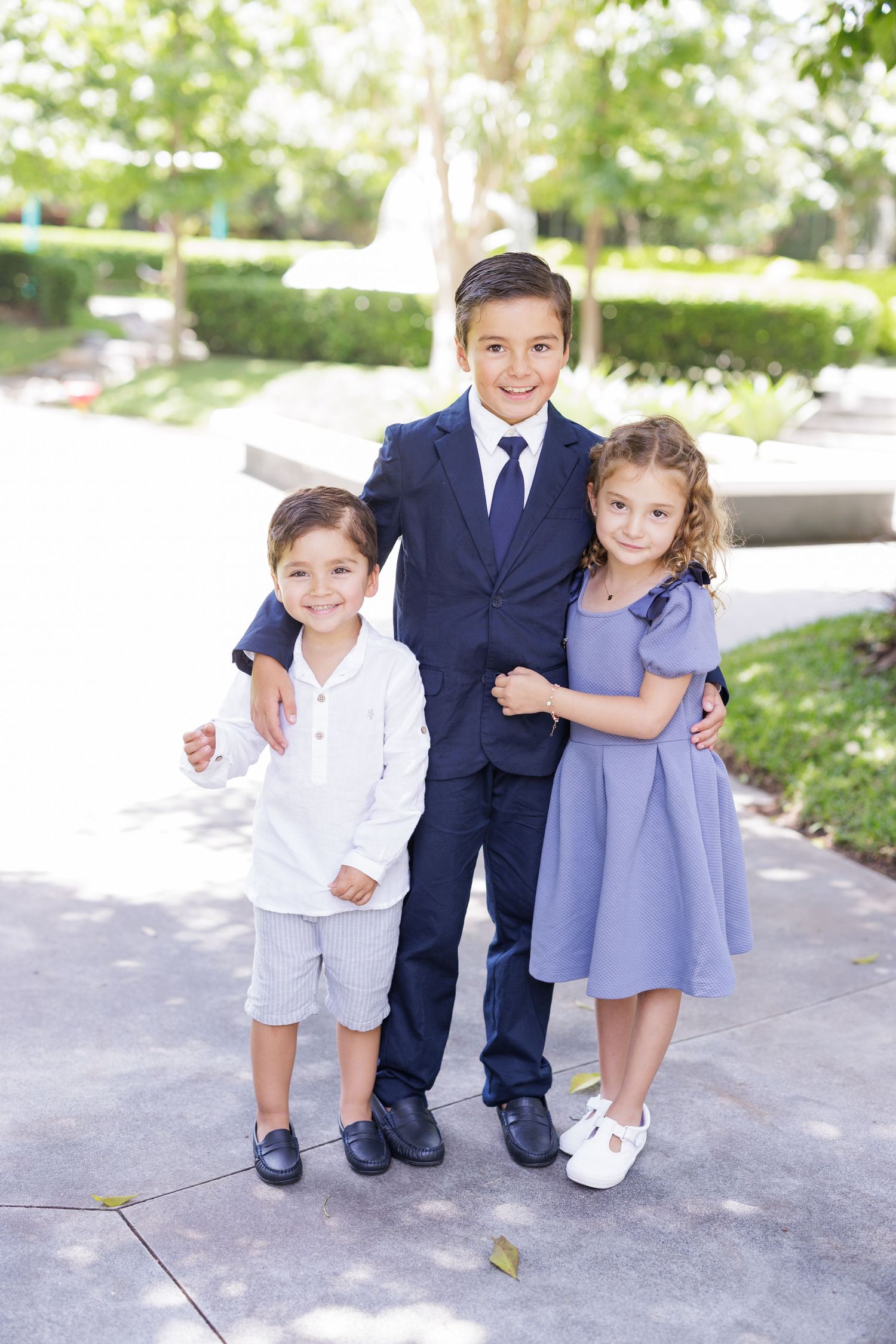 Three well-dressed children pose together outdoors, with the oldest boy in a navy suit flanked by two younger siblings.