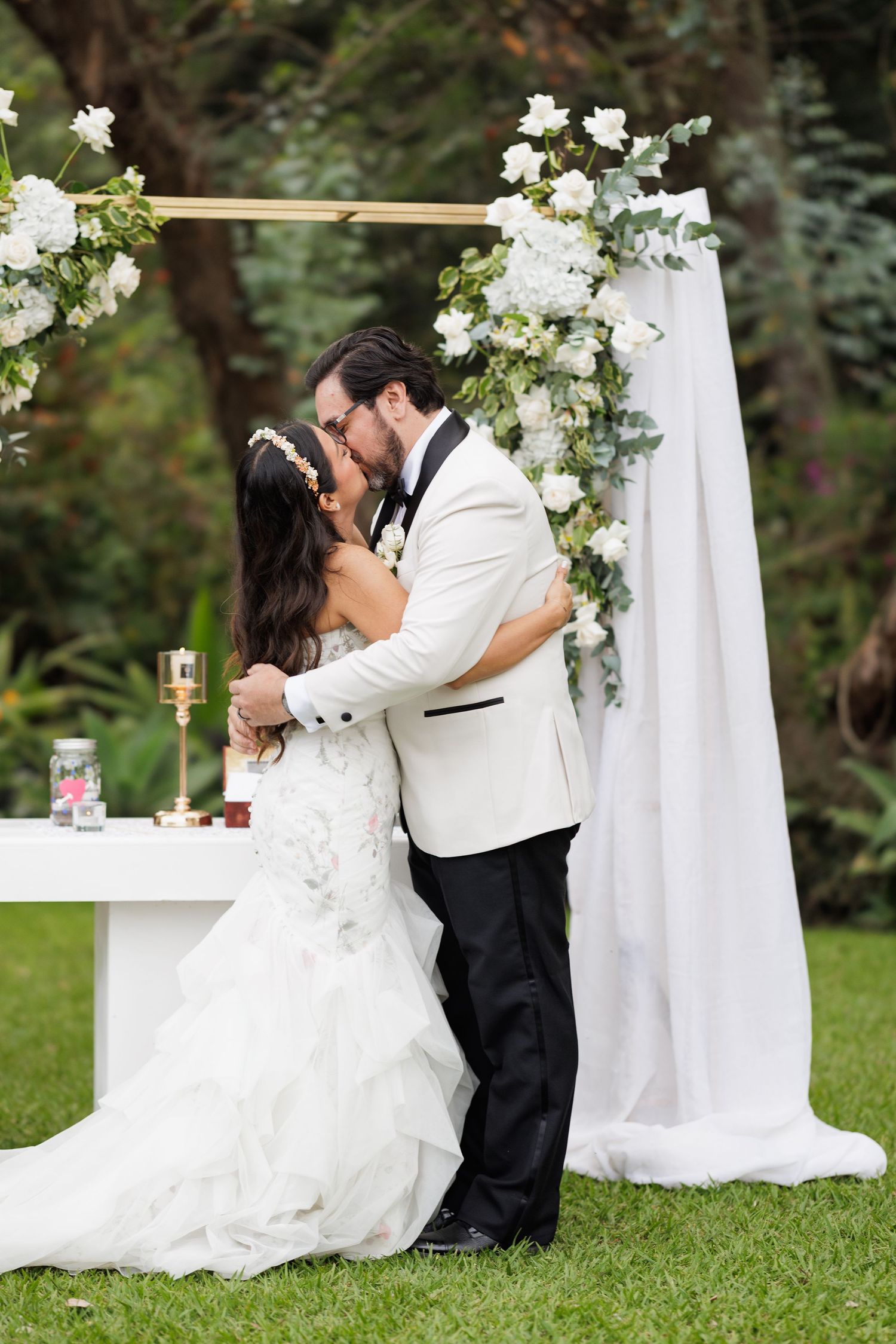 Romantic wedding couple embraces near white floral decorations during their outdoor celebration.
