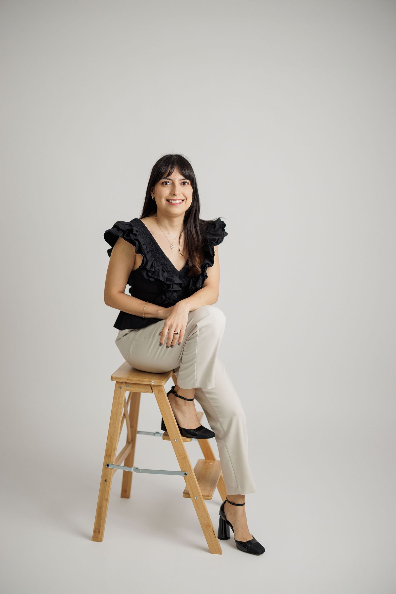 Stylish portraits on wooden stool featuring black ruffled top and light colored pants.