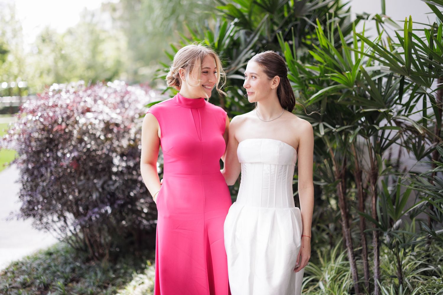 Wedding party members in pink and white dresses gather outdoors in a garden setting.